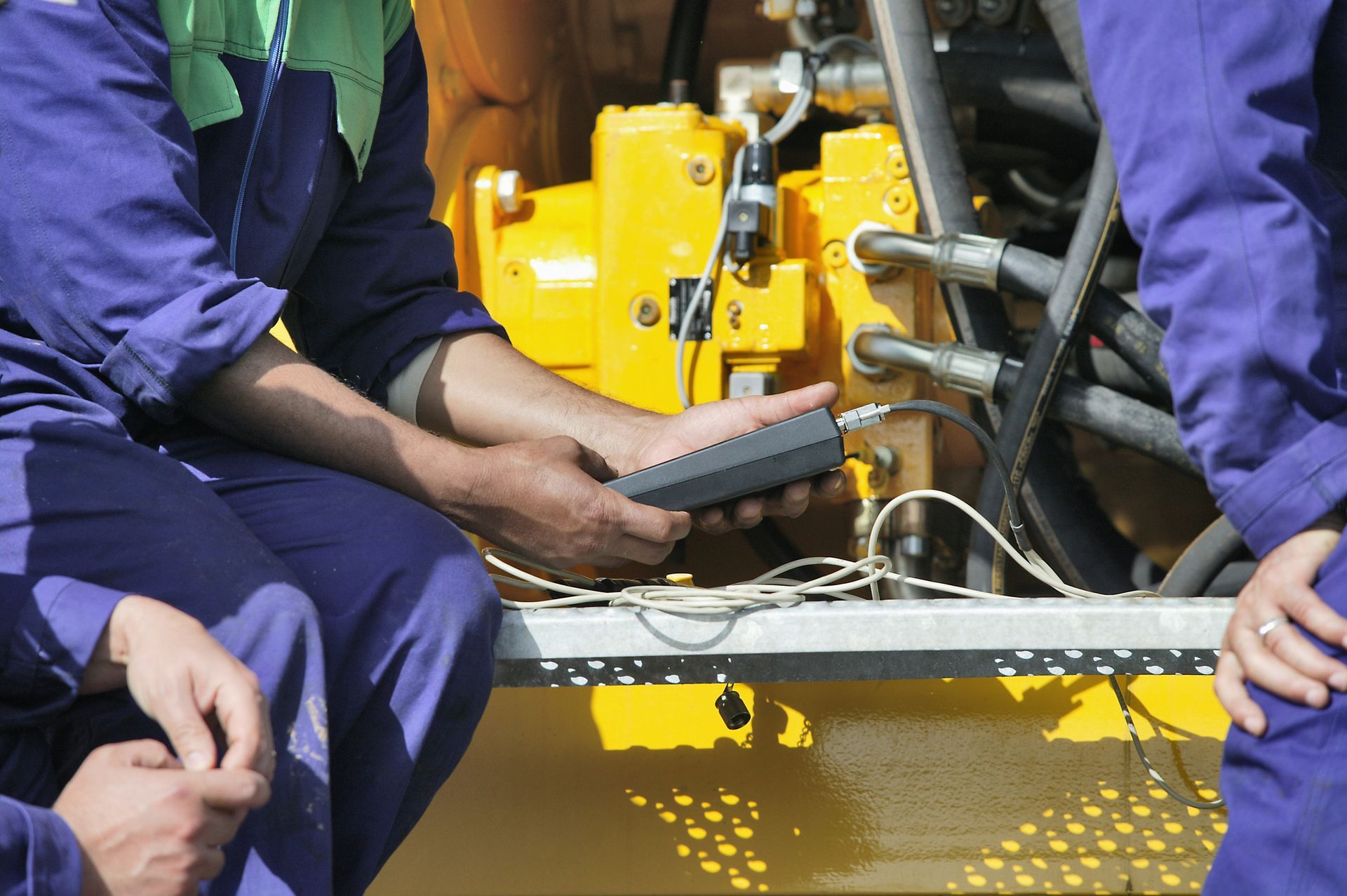 Workers in blue coveralls examining machinery with a handheld device, possibly for maintenance or diagnostics.