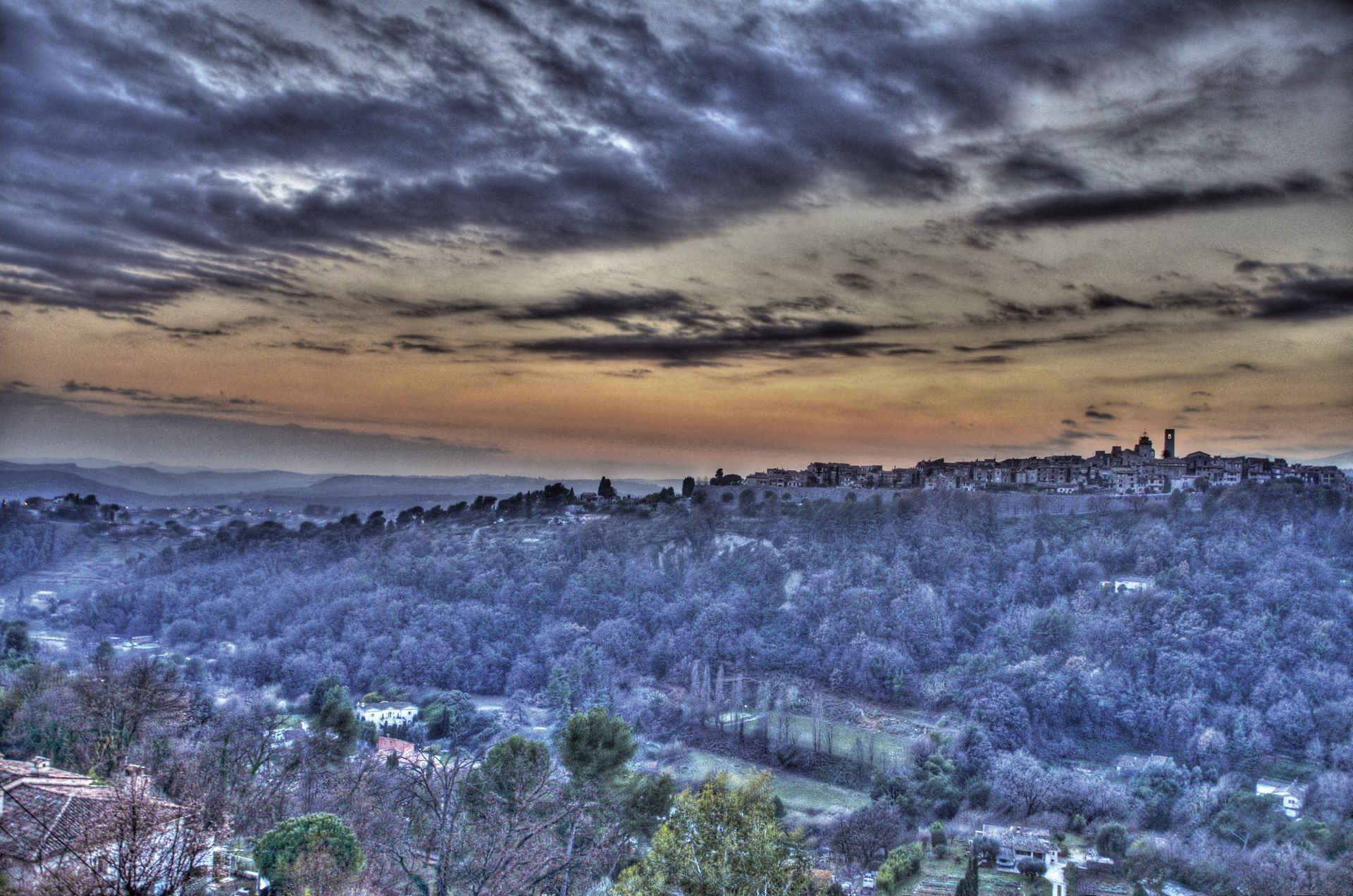 Crépuscule nuageux sur une colline boisée aux tons bleutés et la silhouette lointaine d'une ville