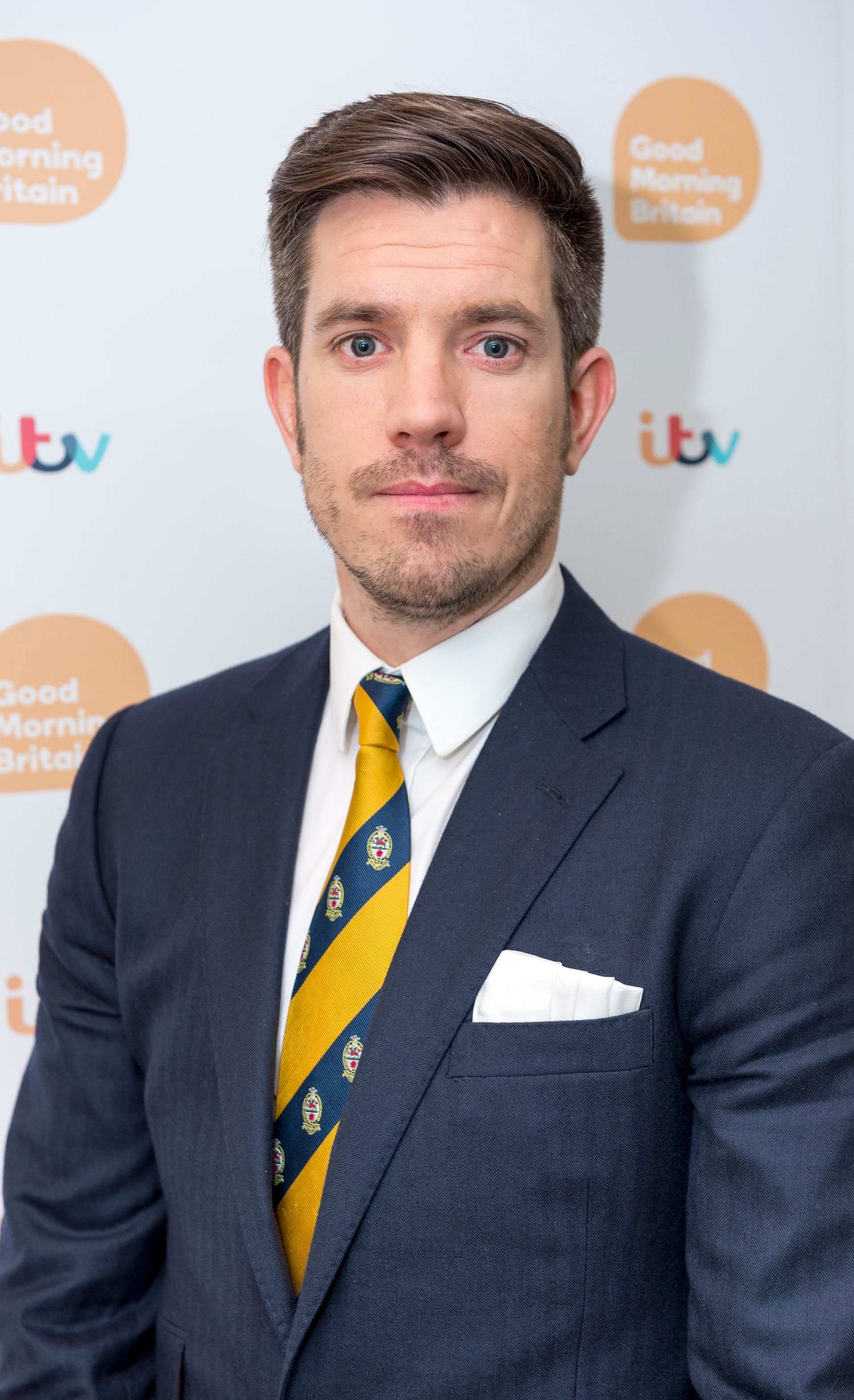 A man in a suit and tie is standing in front of a good morning britain wall.