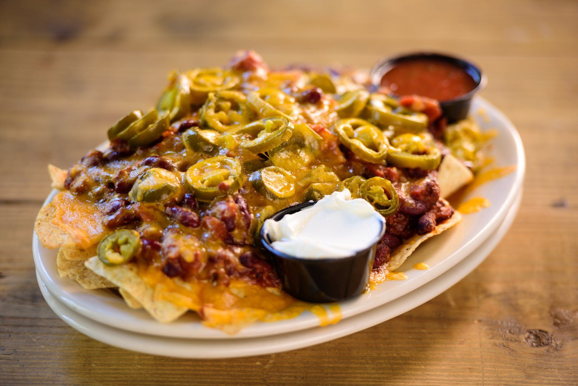 Nachos loaded with cheese, chili, jalapeƱos, and toppings, served with salsa and sour cream on a white plate.