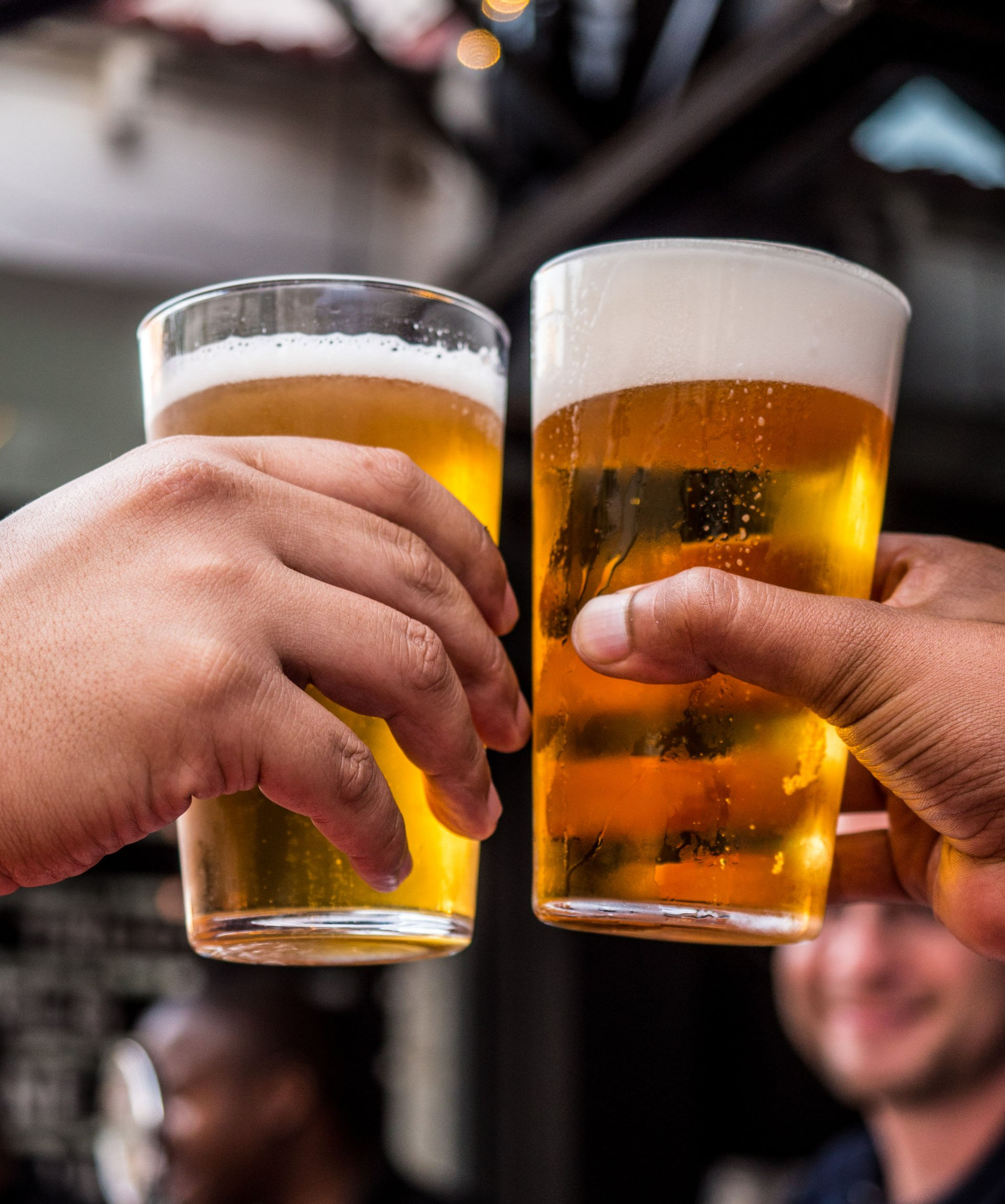 Two hands clink beer glasses in a toast, filled with golden beer and white foam. Outdoor setting with people.