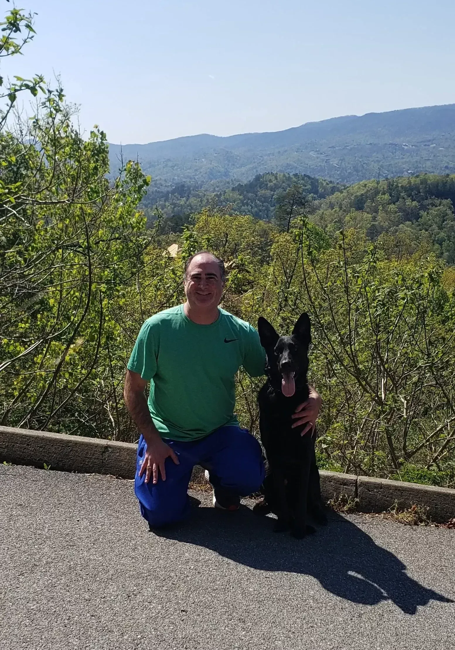 A man in a green shirt is kneeling down next to a black dog.