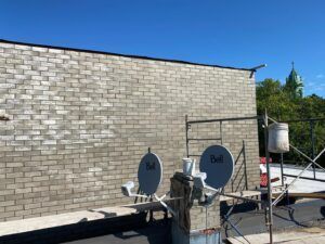 Deux antennes paraboliques sur un toit, face à un mur de briques et un ciel bleu clair. On aperçoit le clocher d'une église au loin.