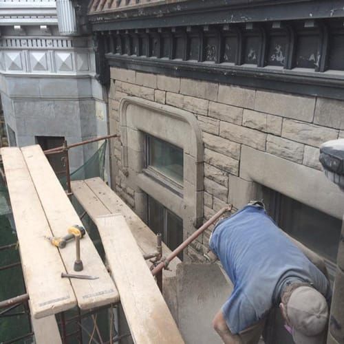 Un ouvrier du bâtiment sur un échafaudage travaille à l'extérieur d'un bâtiment en pierre. Des outils et des planches de bois sont posés sur l'échafaudage.