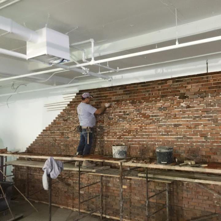 Un ouvrier du bâtiment répare un mur de briques debout sur un échafaudage. Le mur est composé d'un mélange de briques anciennes et neuves.