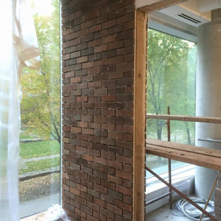 Mur de briques en construction à côté d'une fenêtre, avec vue sur des arbres et un bâtiment sous échafaudage.