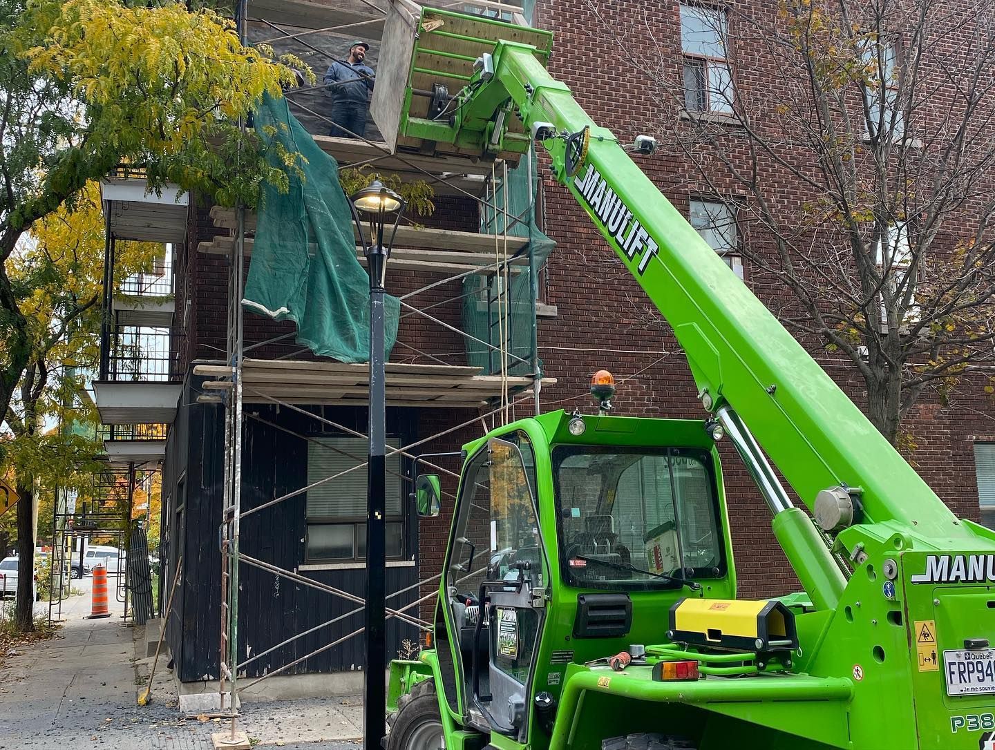 Un chariot télescopique vert soulève des matériaux près de la façade d'un bâtiment avec un échafaudage. Des ouvriers sont visibles près du sommet.