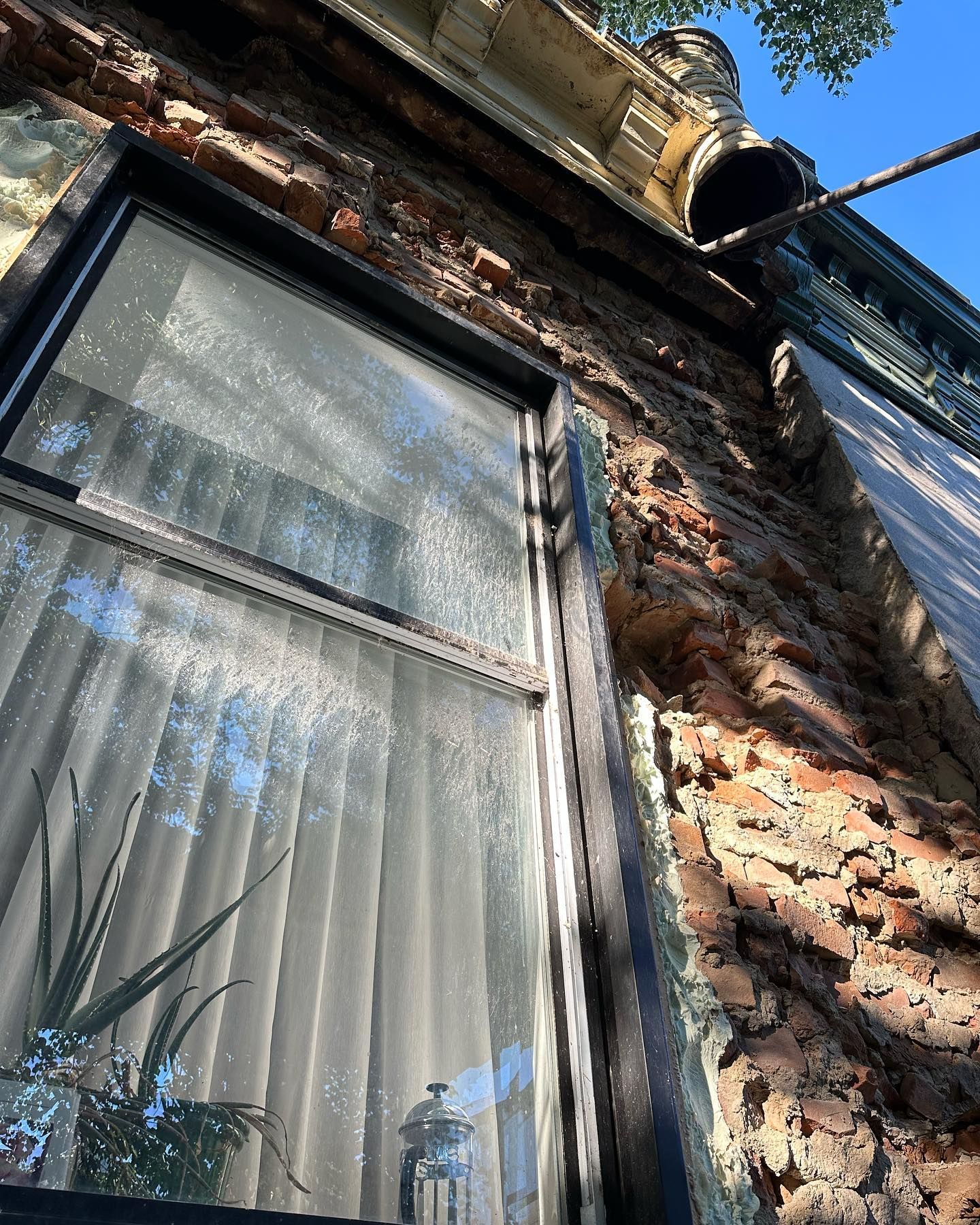 Un bâtiment en briques patinées par le temps avec une fenêtre, vu en contre-plongée. La lumière du soleil se reflète sur la vitre et illumine un rideau.