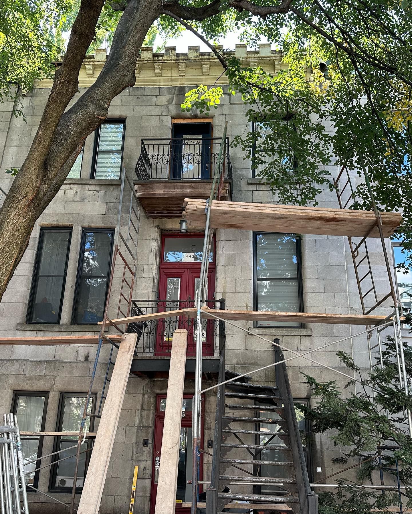 Bâtiment en pierre avec une porte d'entrée rouge, un escalier de secours en métal noir et des échafaudages, suggérant une rénovation. Un arbre ombrage les étages supérieurs.