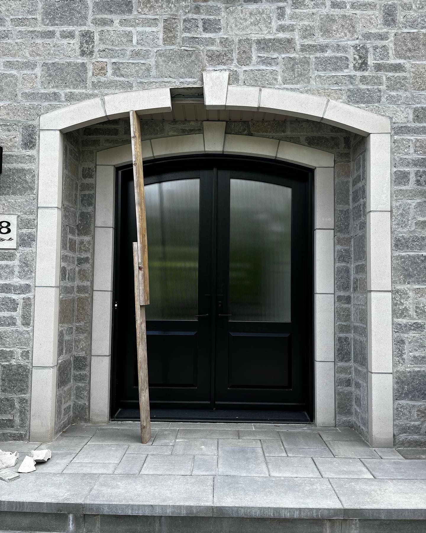 Portes doubles noires avec fenêtres cintrées, arche et moulures en pierre claire. Un support en bois est placé devant l'une des portes.