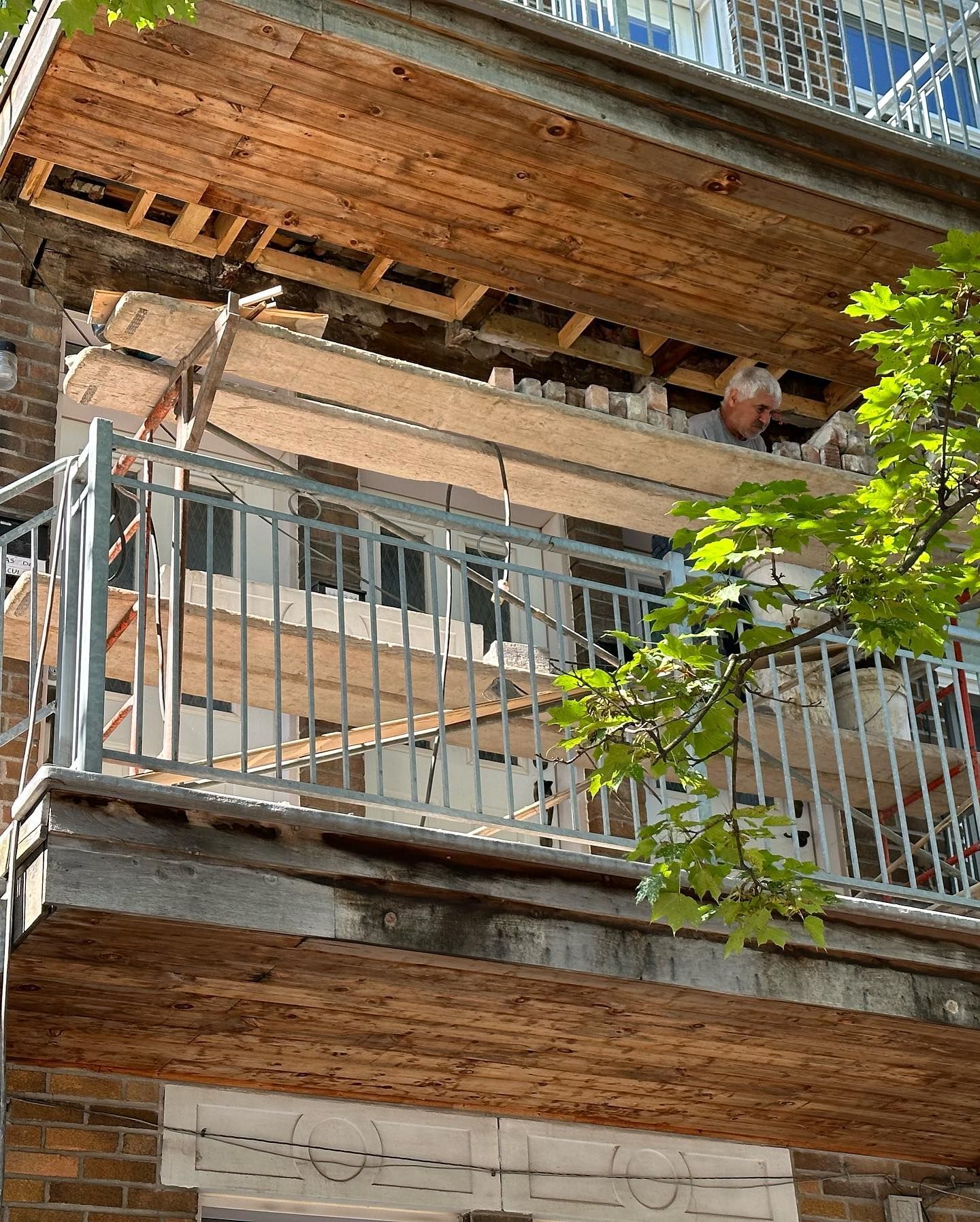 Balcon en construction sur un immeuble en briques. Des planches de bois et un échafaudage sont visibles. Une personne travaille sur le balcon.