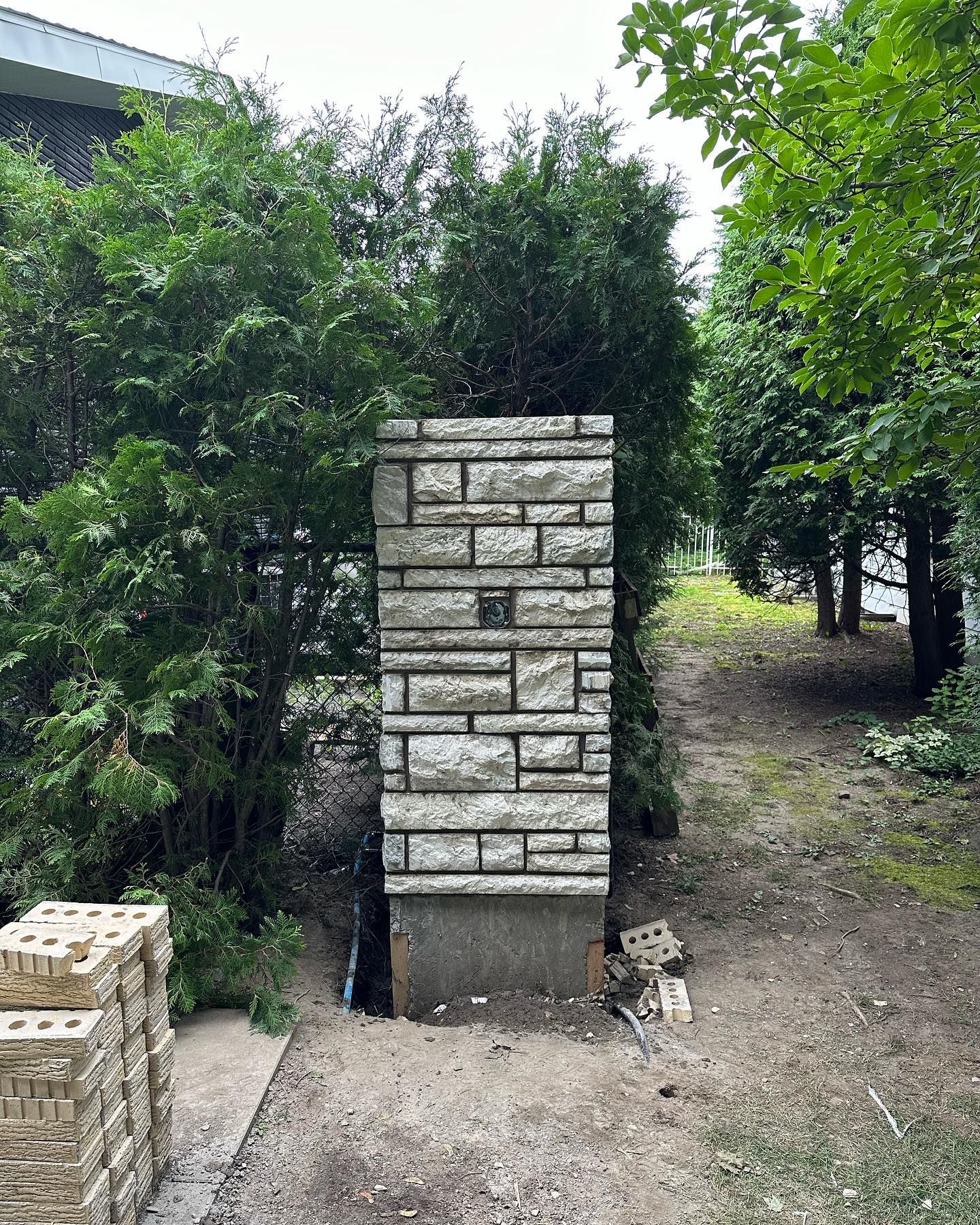 Pilier de pierre devant des arbustes et des arbres verts, marquant peut-être une entrée. Des briques sont empilées à gauche.