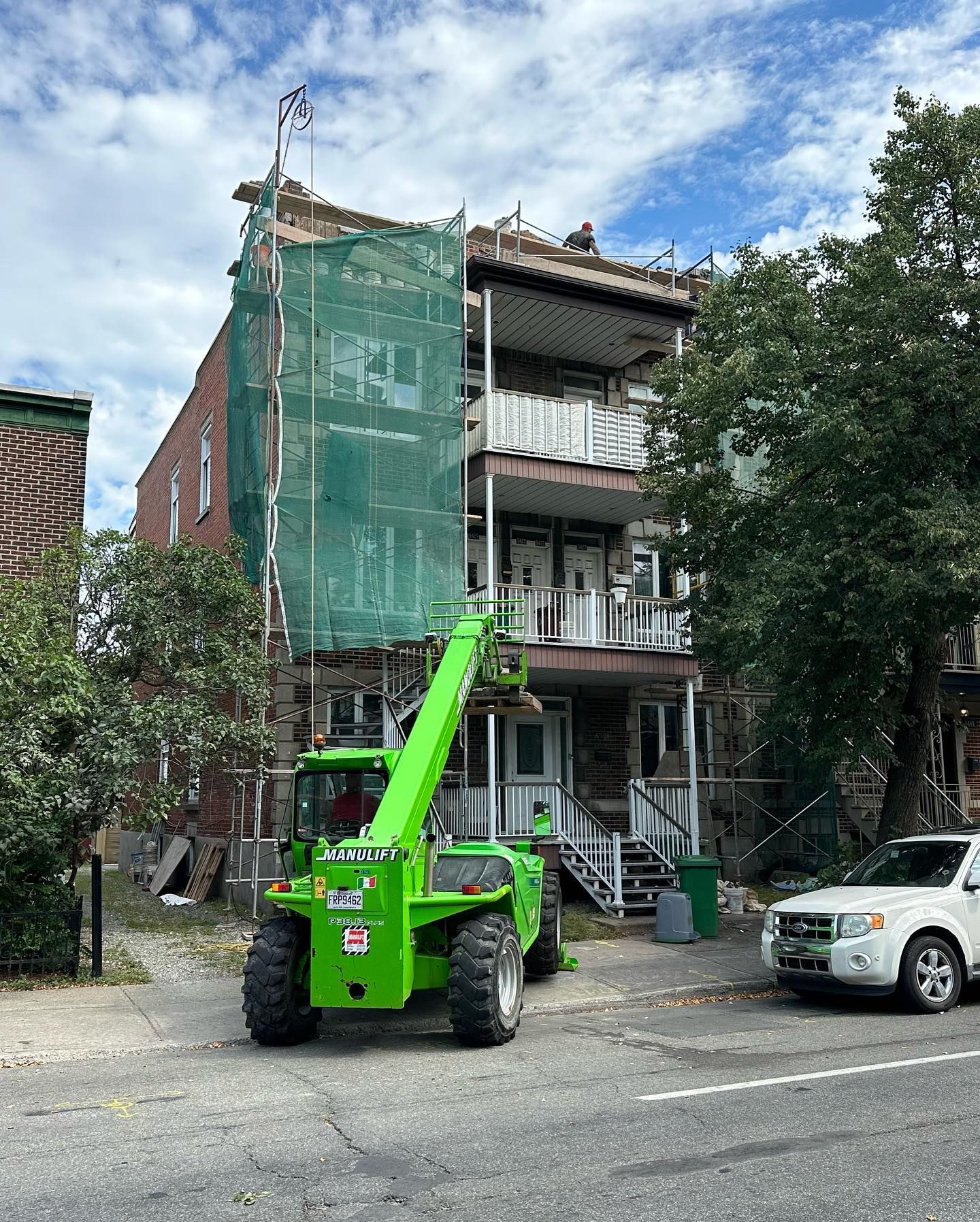 Un chariot élévateur vert à côté d'un bâtiment en construction ; un filet vert recouvre le côté. Un SUV blanc est garé dans la rue.