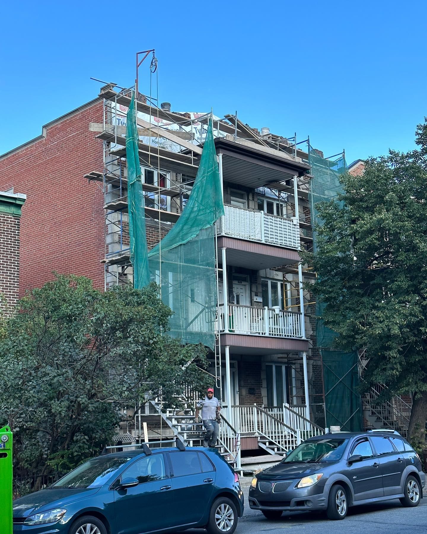 Bâtiment en construction avec échafaudages et filets verts. Des voitures sont garées dans la rue devant.