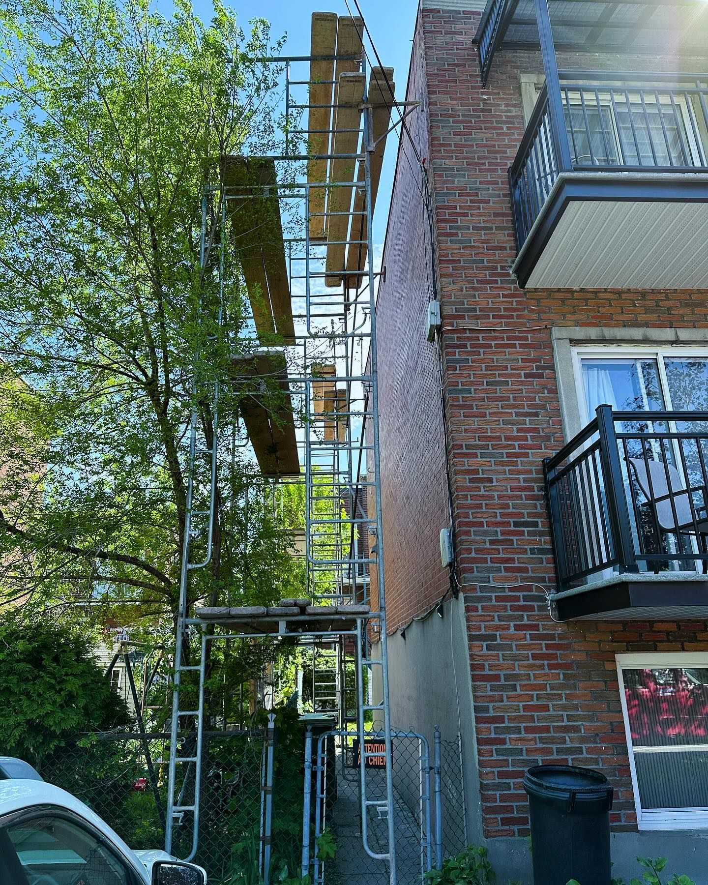 Échafaudage à côté d'un immeuble en briques. Des planches de bois forment des plateformes ; un arbre et une poubelle noire sont également présents.