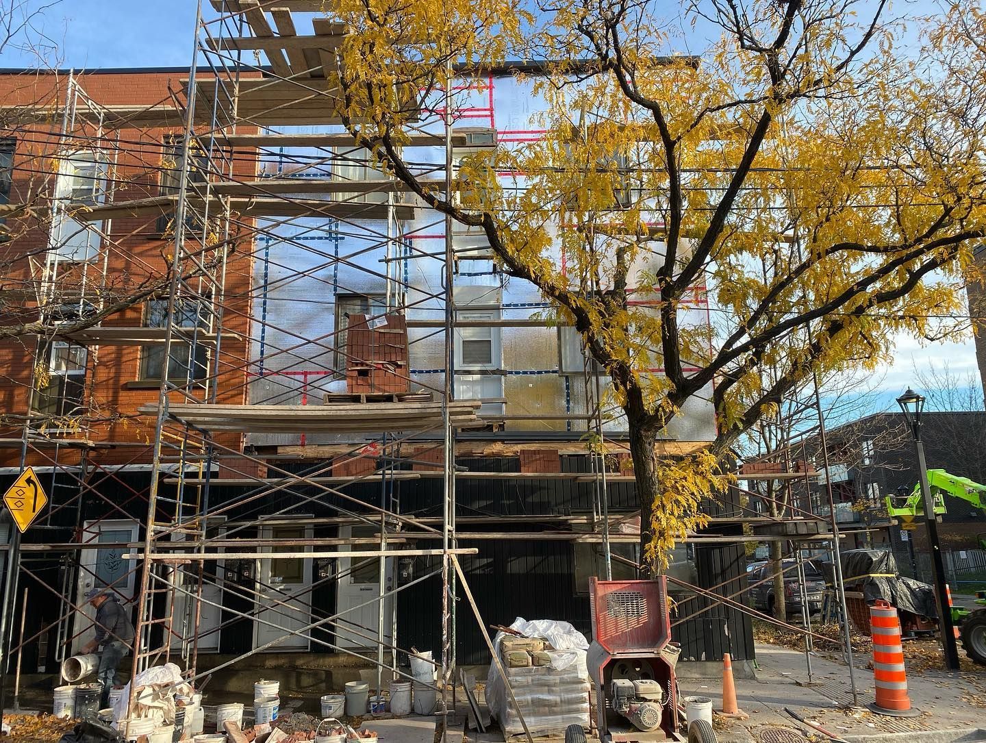 Bâtiment en construction avec échafaudages, façade en briques et isolation réfléchissante visibles. Un arbre aux feuilles jaunes se dresse devant.