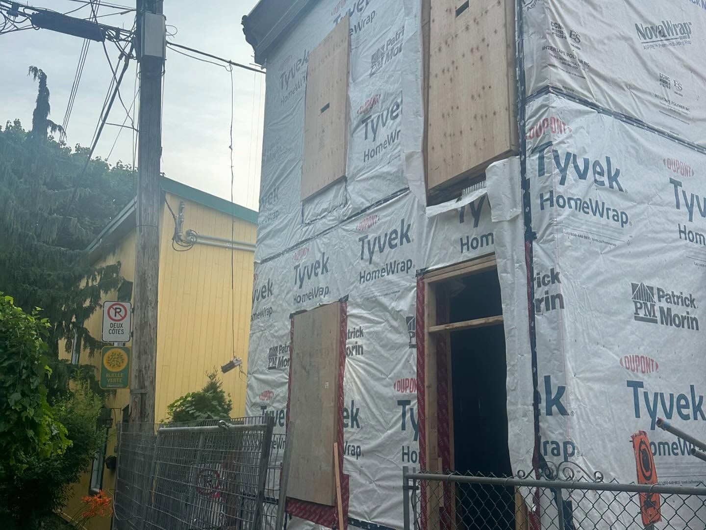 Maison en rénovation. Extérieur recouvert de Tyvek, fenêtres bardées, poteau électrique à côté. Bâtiment jaune en arrière-plan.