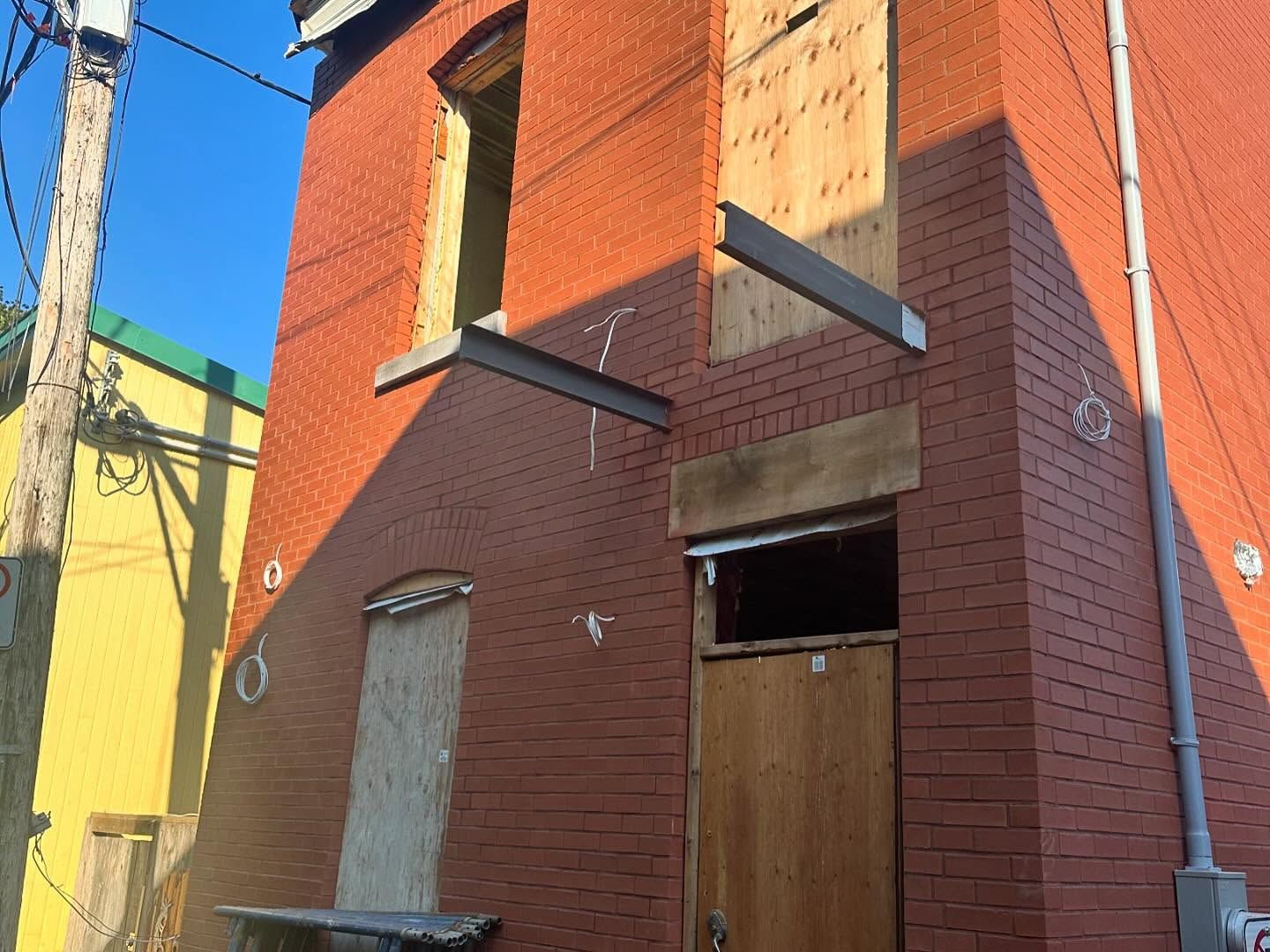 Bâtiment en briques rouges avec fenêtres et porte condamnées. Deux poutres métalliques dépassent du mur, soutenant peut-être quelque chose.
