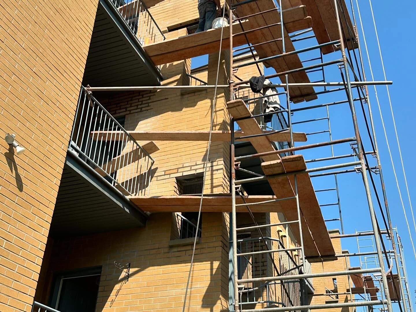 Échafaudage sur un bâtiment en briques avec ouvriers sur les plateformes. Journée ensoleillée avec un balcon visible.