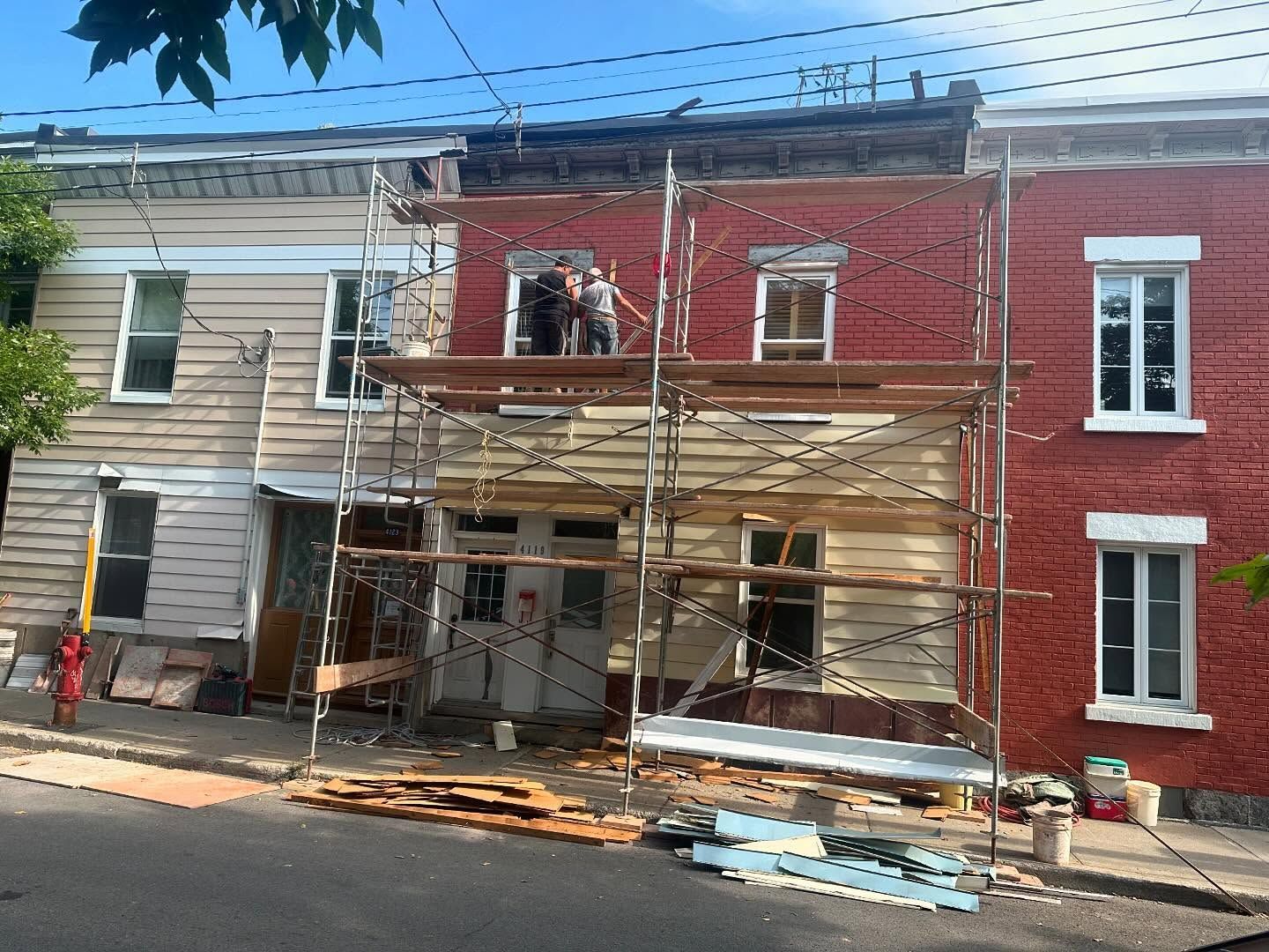 Rangée de maisons, dont une en construction avec échafaudage. Ouvriers sur l'échafaudage, avec revêtement rouge et crème.