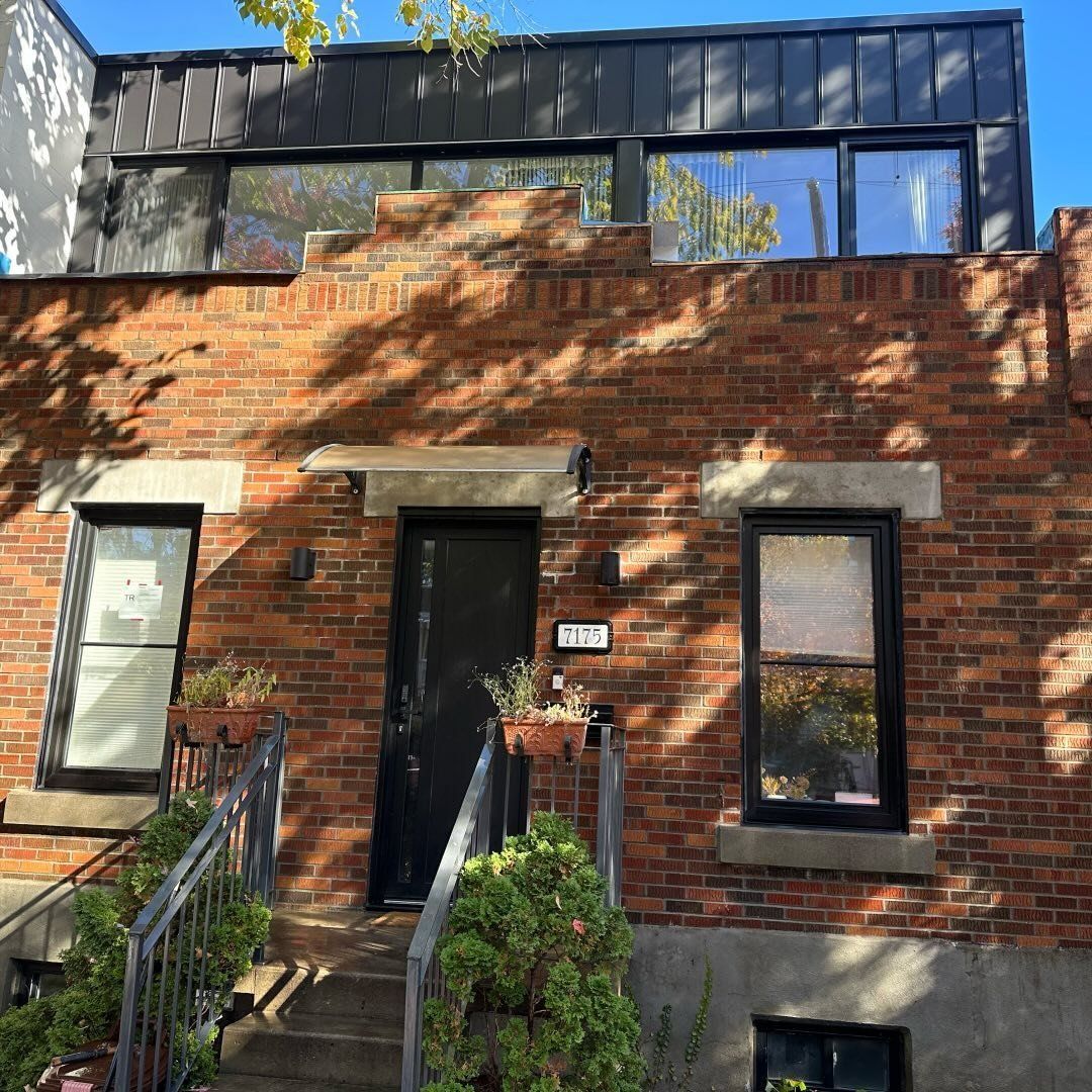 Maison en briques rouges avec porte noire et étage en métal foncé. Une végétation luxuriante encadre l'entrée.