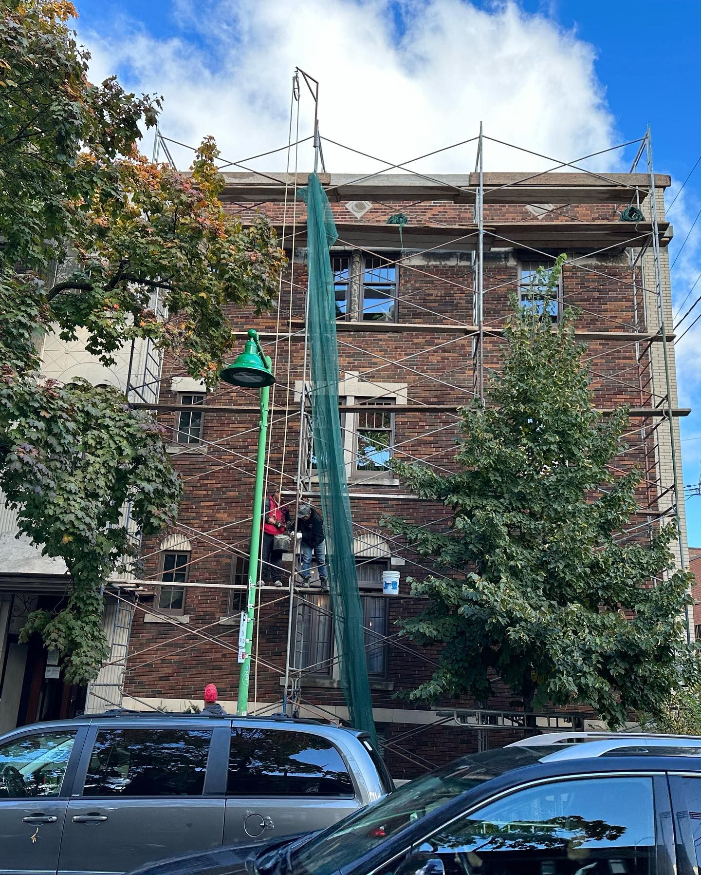 Bâtiment en briques en cours de rénovation, dissimulé par des échafaudages et des filets verts. Voitures garées dans la rue devant.