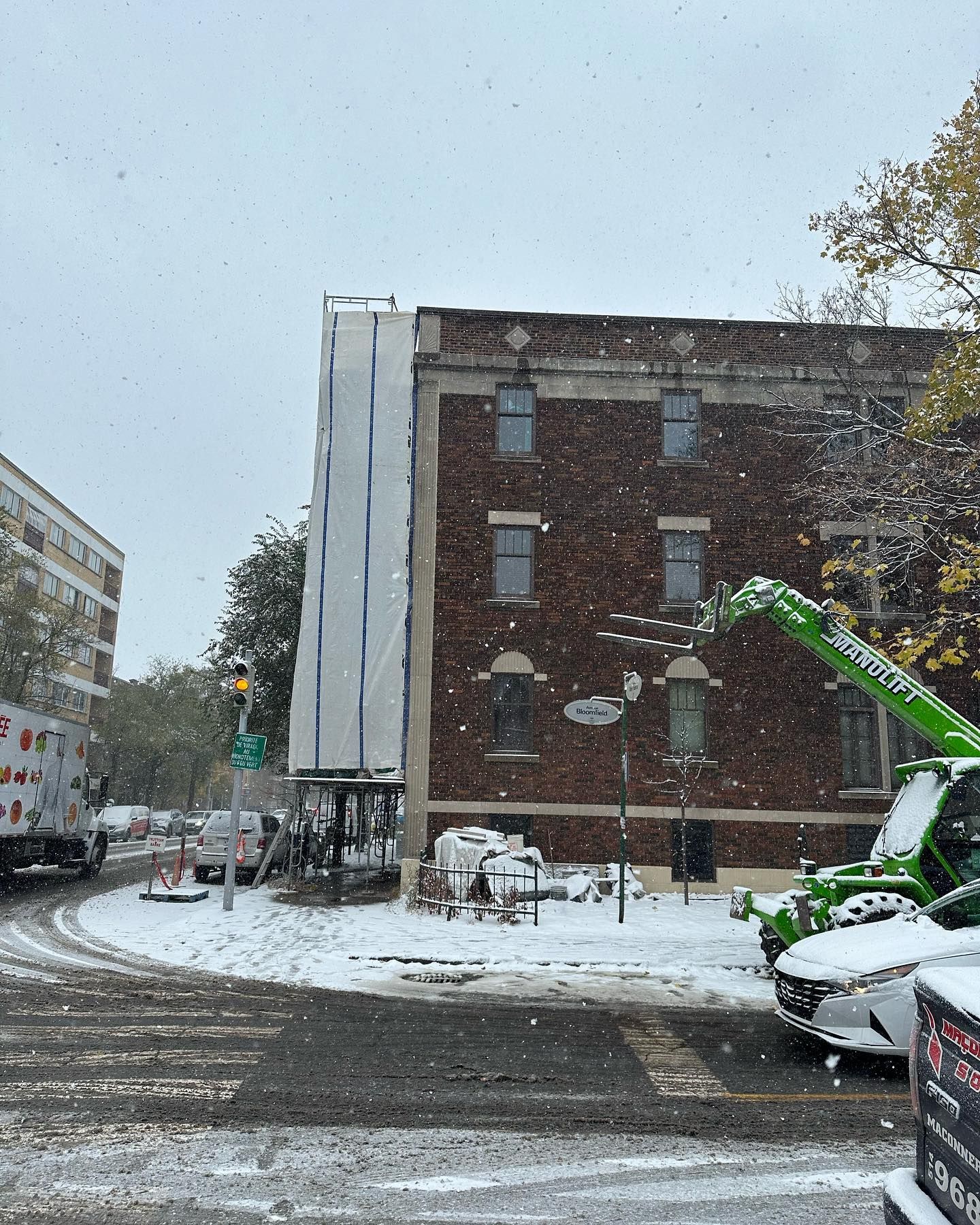 Scène de rue enneigée avec un bâtiment en briques partiellement recouvert d'une bâche blanche pour réparations. Une nacelle élévatrice verte se trouve à proximité.