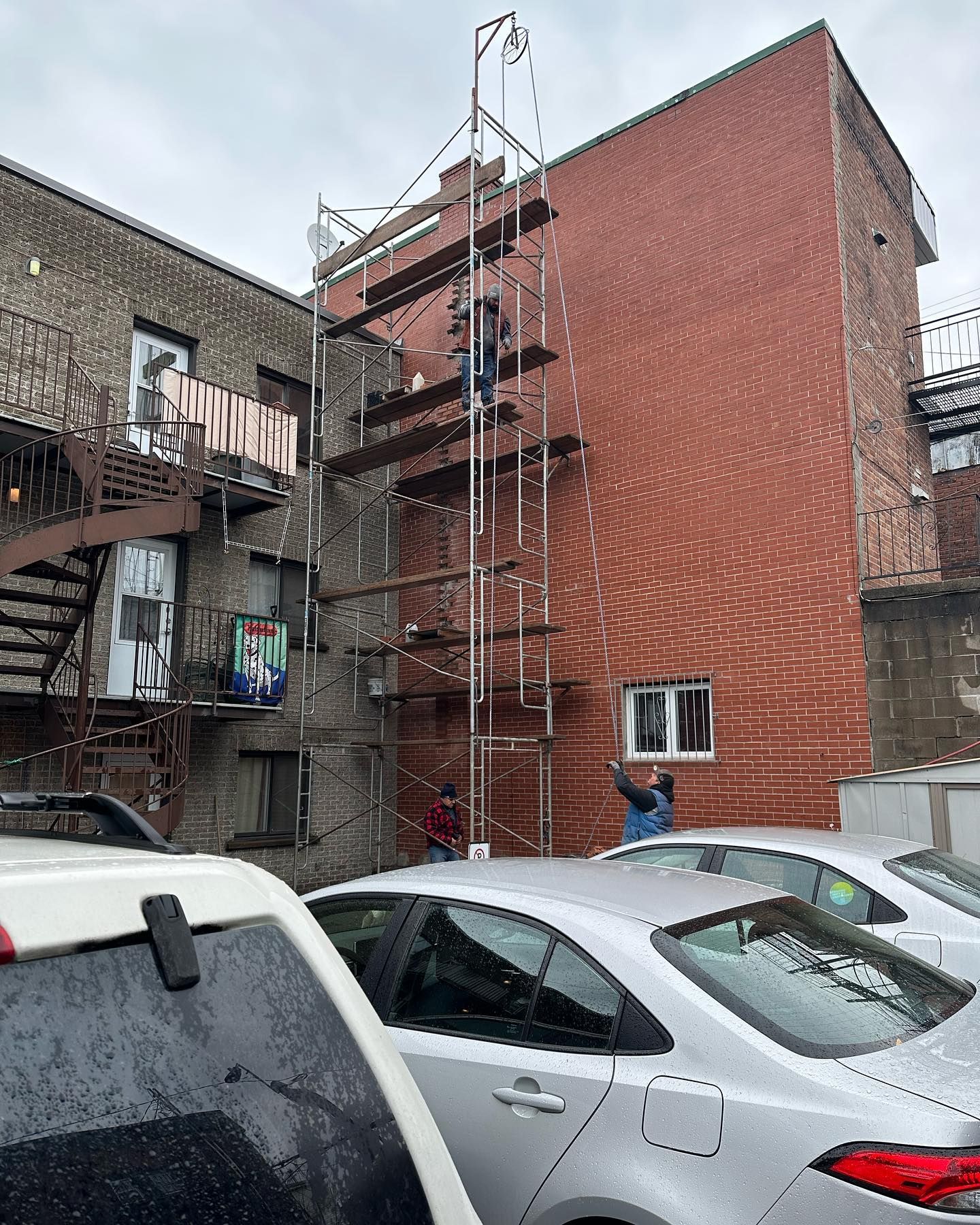 Échafaudage sur un bâtiment en briques avec ouvriers. Des voitures grises sont garées au premier plan.