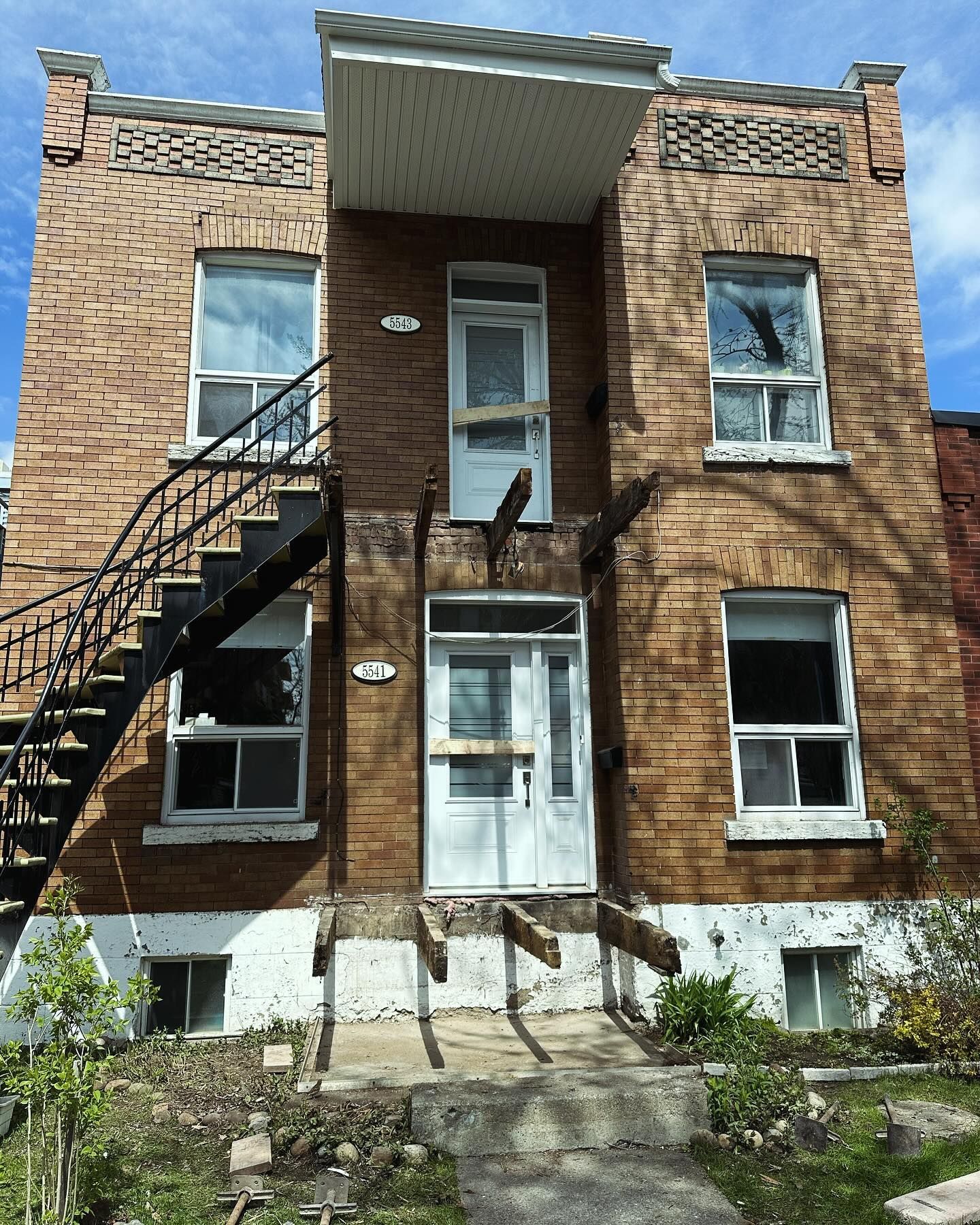 Bâtiment en briques de deux étages avec escalier extérieur en métal, deux portes blanches et supports en bois cassés à l'entrée.