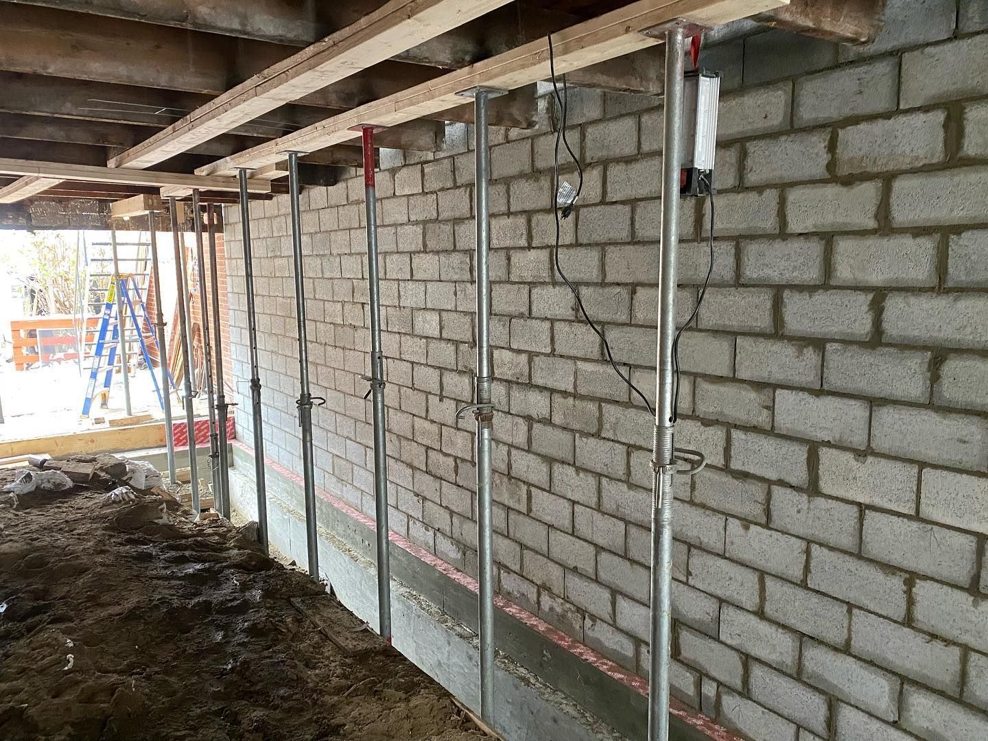Chantier de construction avec des vérins de support métalliques soutenant un mur de briques sous des poutres en bois.