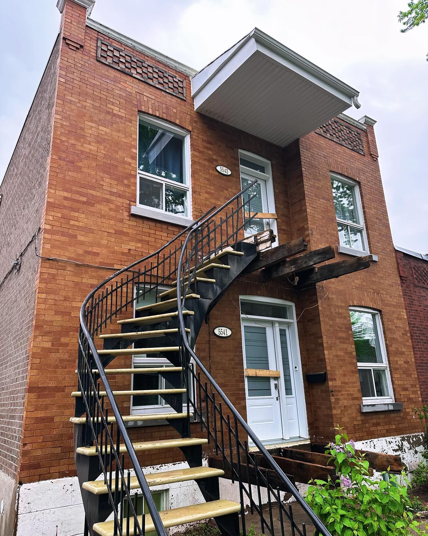 Immeuble en briques avec escalier extérieur en métal noir. Portes et fenêtres blanches sur plusieurs niveaux.