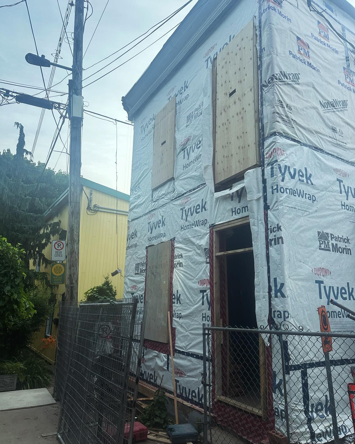 Un bâtiment de deux étages en construction, enveloppé de Tyvek, avec des fenêtres barricadées et un poteau électrique devant.