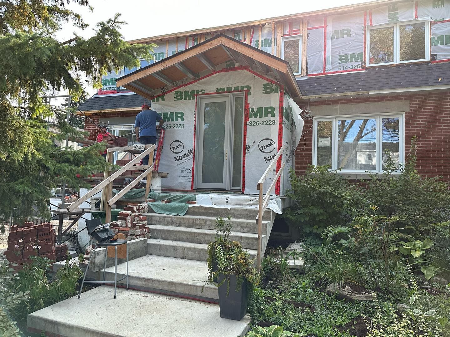 Extérieur de la maison en construction ; porche avec nouveau toit, marches et revêtement en cours d'installation ; personne travaillant sur le toit du porche.