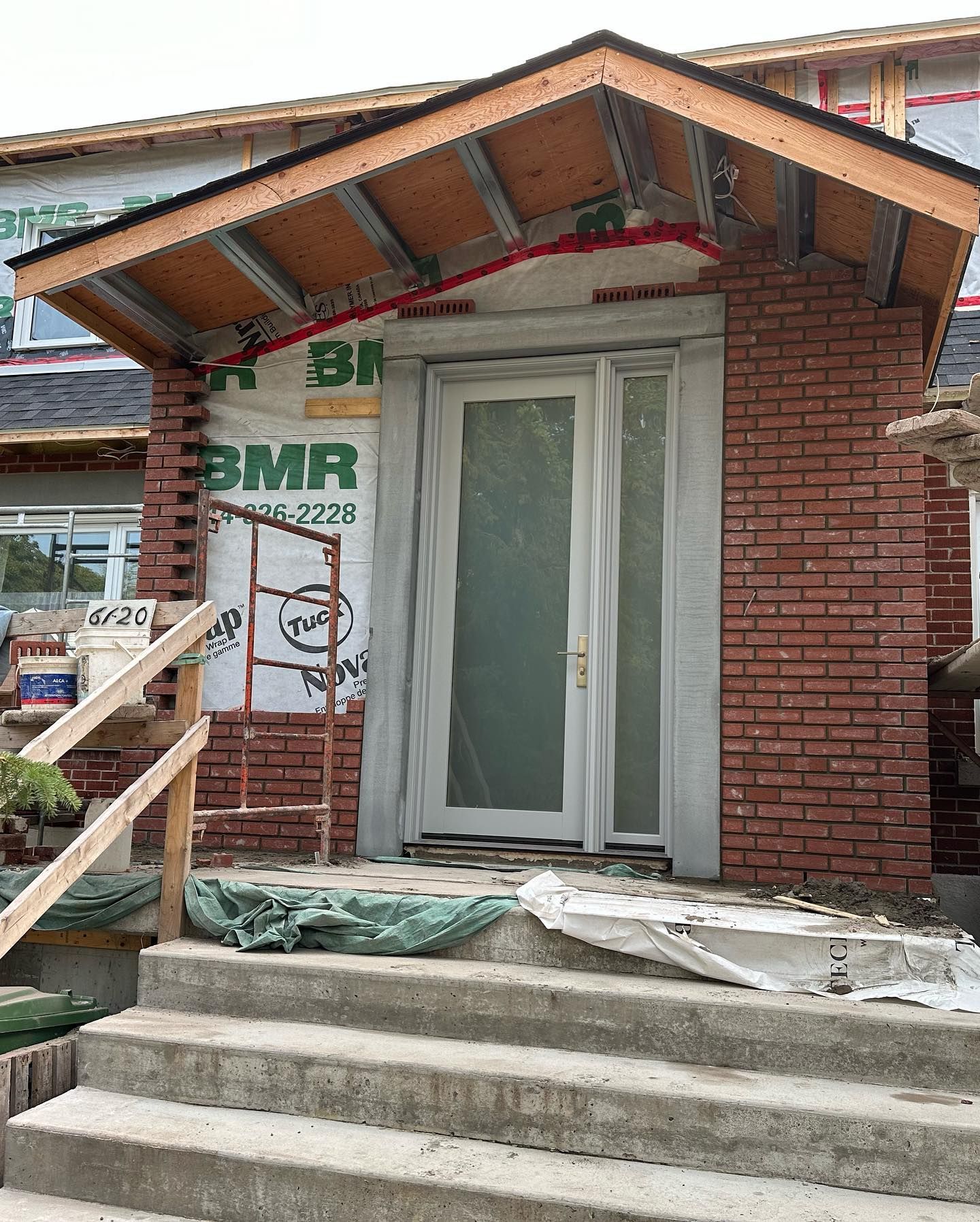 Une entrée aux murs de briques avec une porte blanche sous un toit en bois. Des marches en béton mènent à l'entrée, en cours de construction.