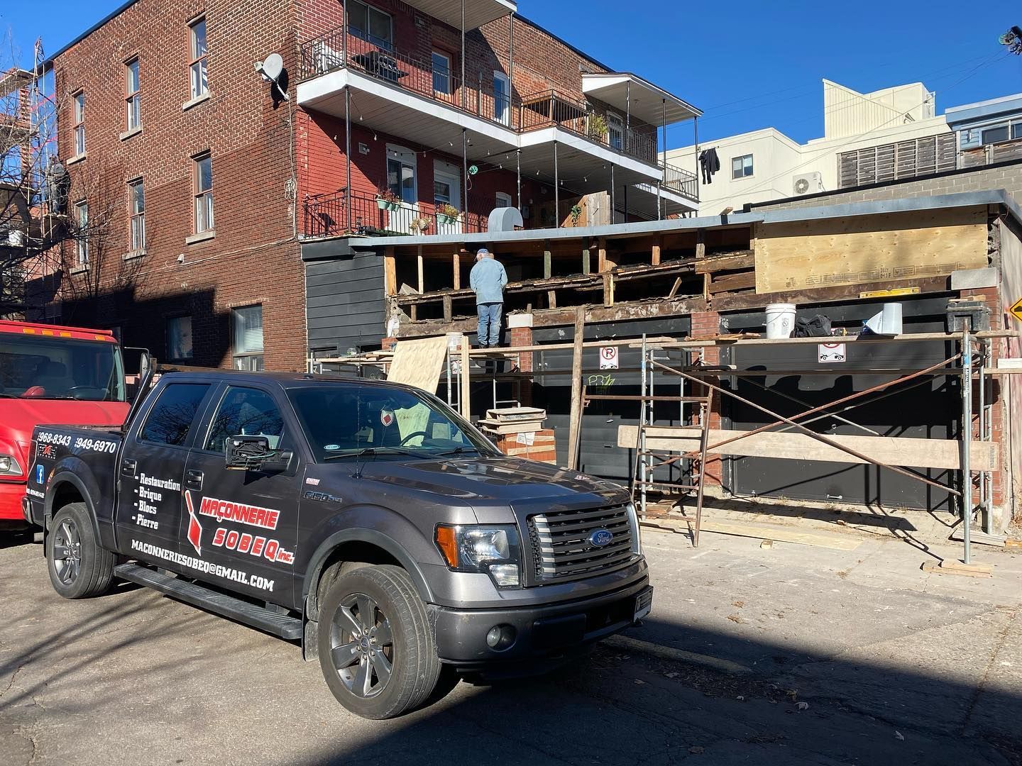 Une camionnette grise est garée devant un immeuble en construction. Deux ouvriers sont visibles sur le toit.