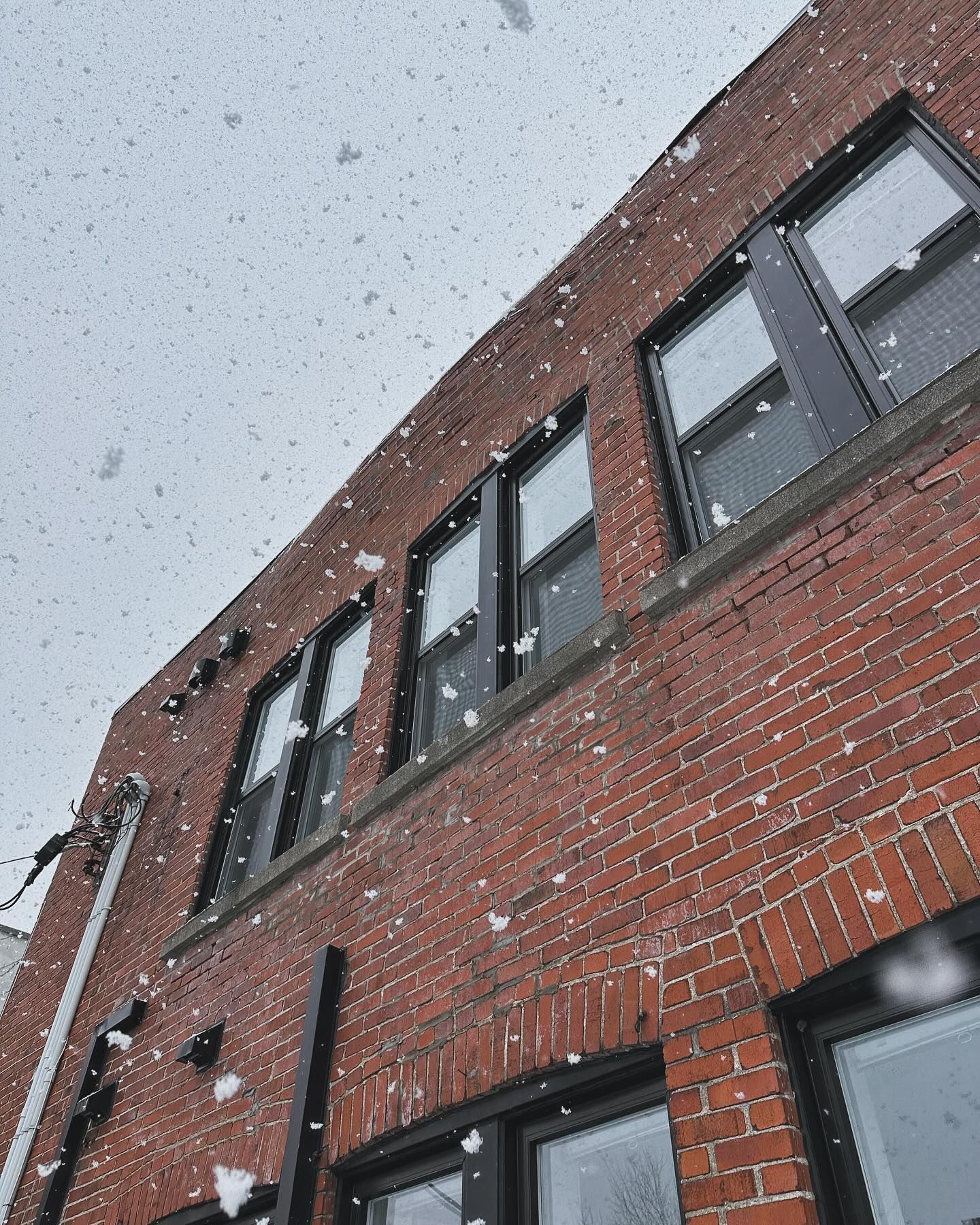 La neige tombe sur un bâtiment en briques rouges aux fenêtres aux cadres noirs. Un ciel gris s'étend au-dessus.