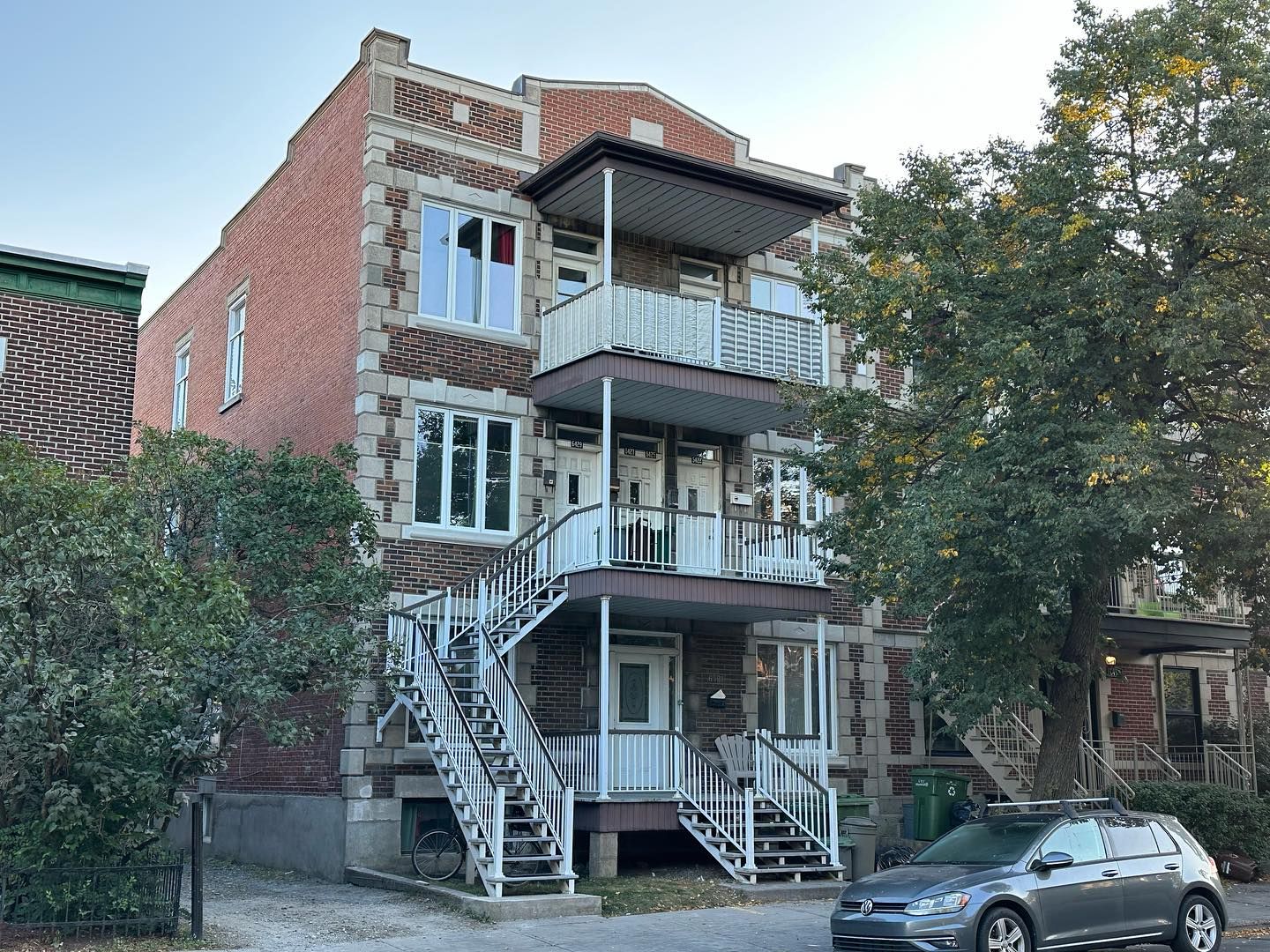 Immeuble en briques à plusieurs étages avec escaliers extérieurs et balcons. Voiture grise garée devant l'immeuble, dans la rue.