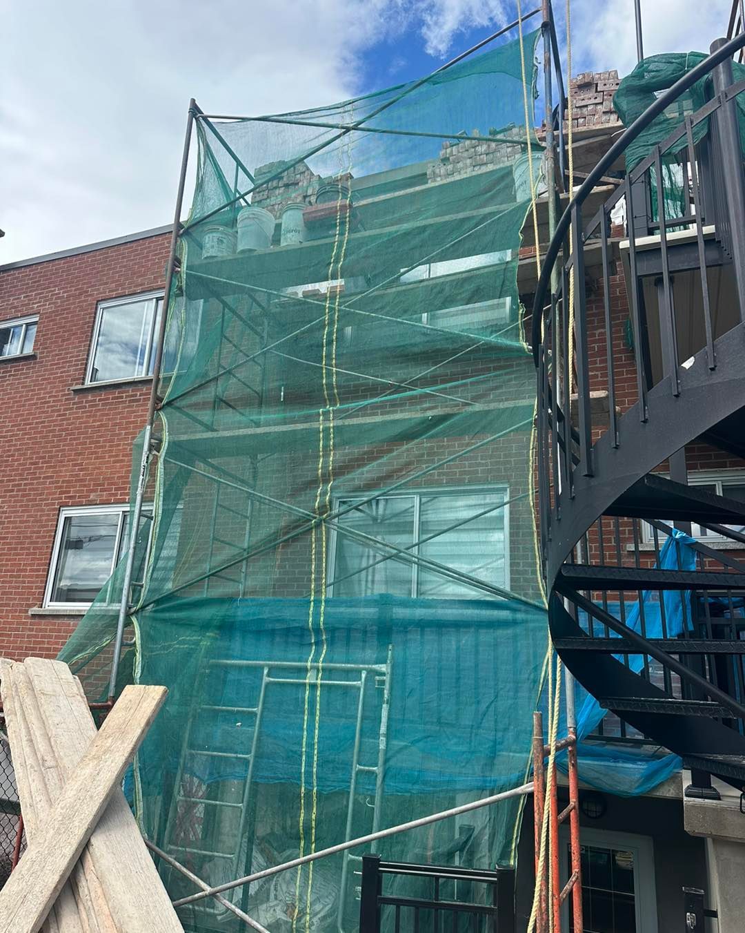 Un échafaudage recouvert d'un filet vert entoure un bâtiment en briques en construction ; un escalier en colimaçon est visible.