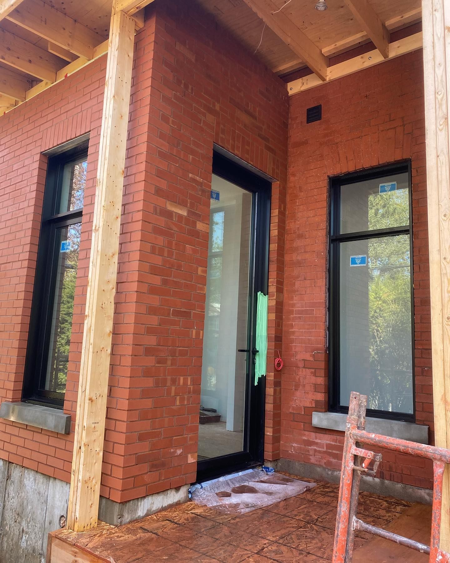 Vue extérieure d'un bâtiment en briques en construction. Il comporte une porte et deux fenêtres à cadre noir. Des poutres en bois et un échafaudage sont également visibles.