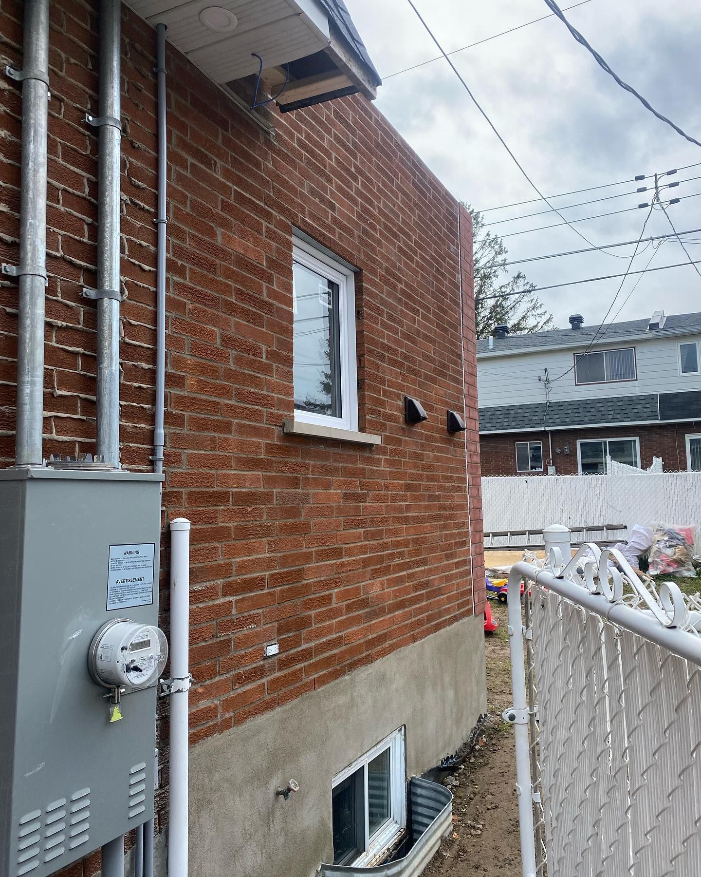 Façade d'un bâtiment en briques rouges avec compteur électrique, fenêtre et canalisations. Une clôture blanche apparaît au premier plan.