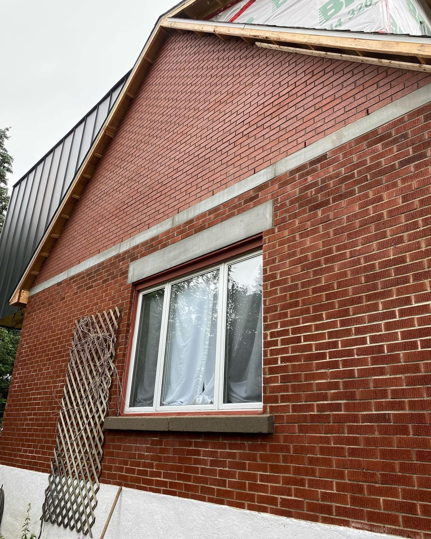 Extérieur d'une maison en briques avec une fenêtre. Façade en briques rouges, encadrement de fenêtre blanc, linteau gris. En construction, charpente apparente.