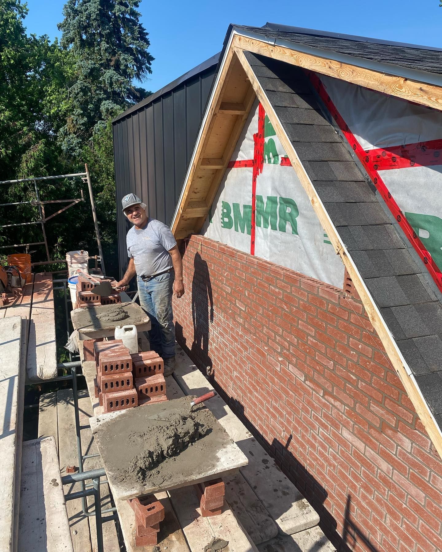 Un homme pose des briques sur le côté d'un bâtiment en construction, vêtu d'une chemise grise et d'un jean. Cadre extérieur ensoleillé.