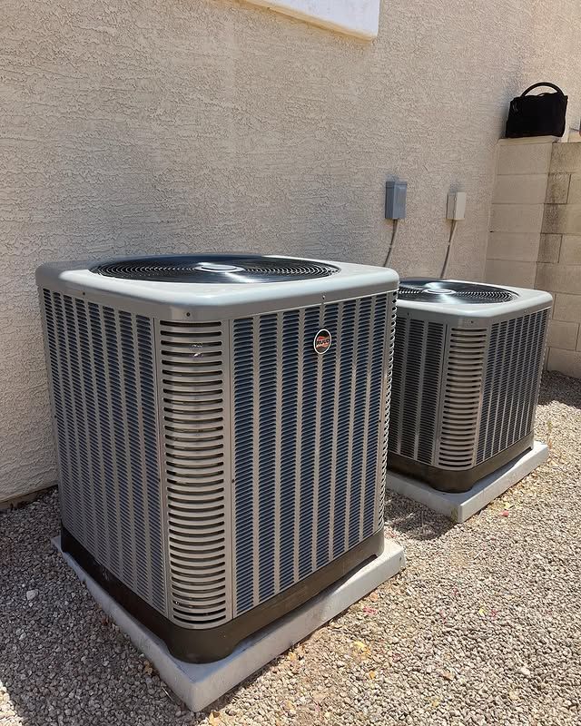 Two gray air conditioning units on concrete pads against a beige wall, gravel ground.
