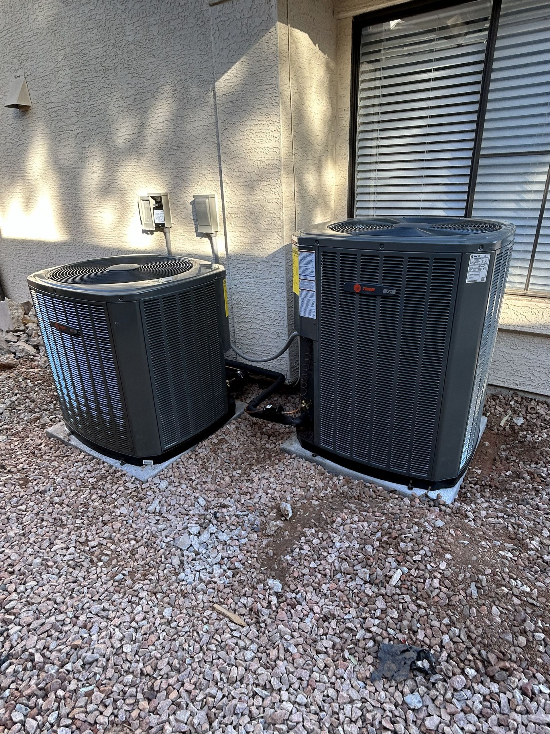 Two black air conditioning units next to a building on a gravel surface.