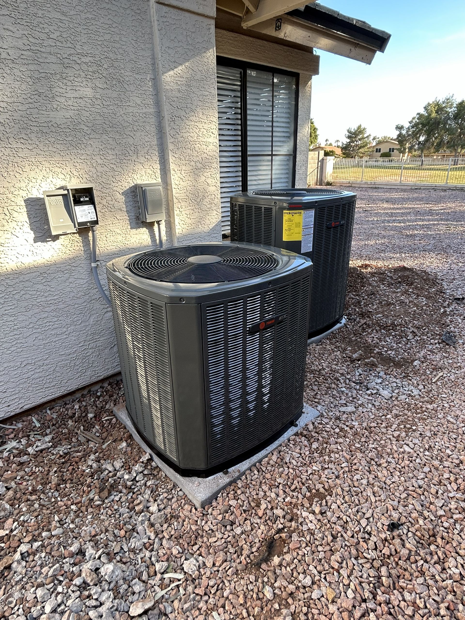 Two dark-colored air conditioning units next to a light-colored stucco building on a gravel surface.