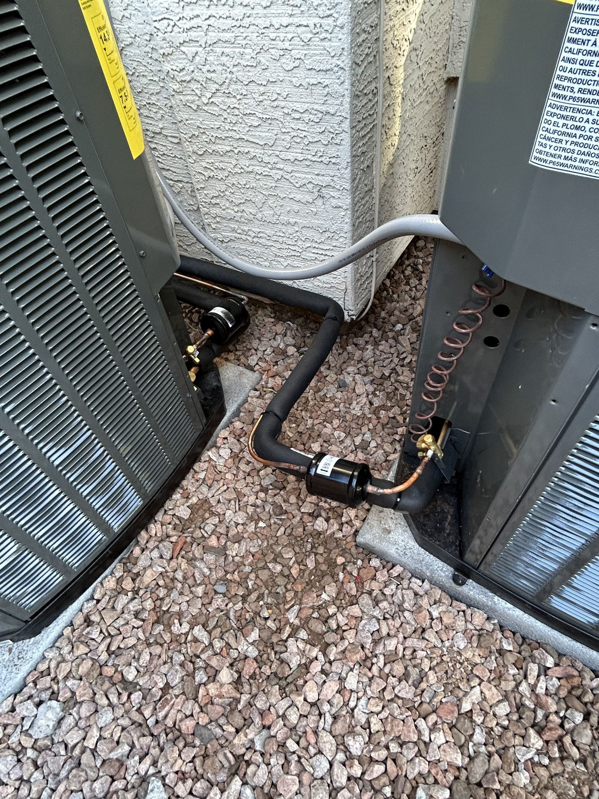 Two air conditioning units connected by black pipes and a filter, on gravel.