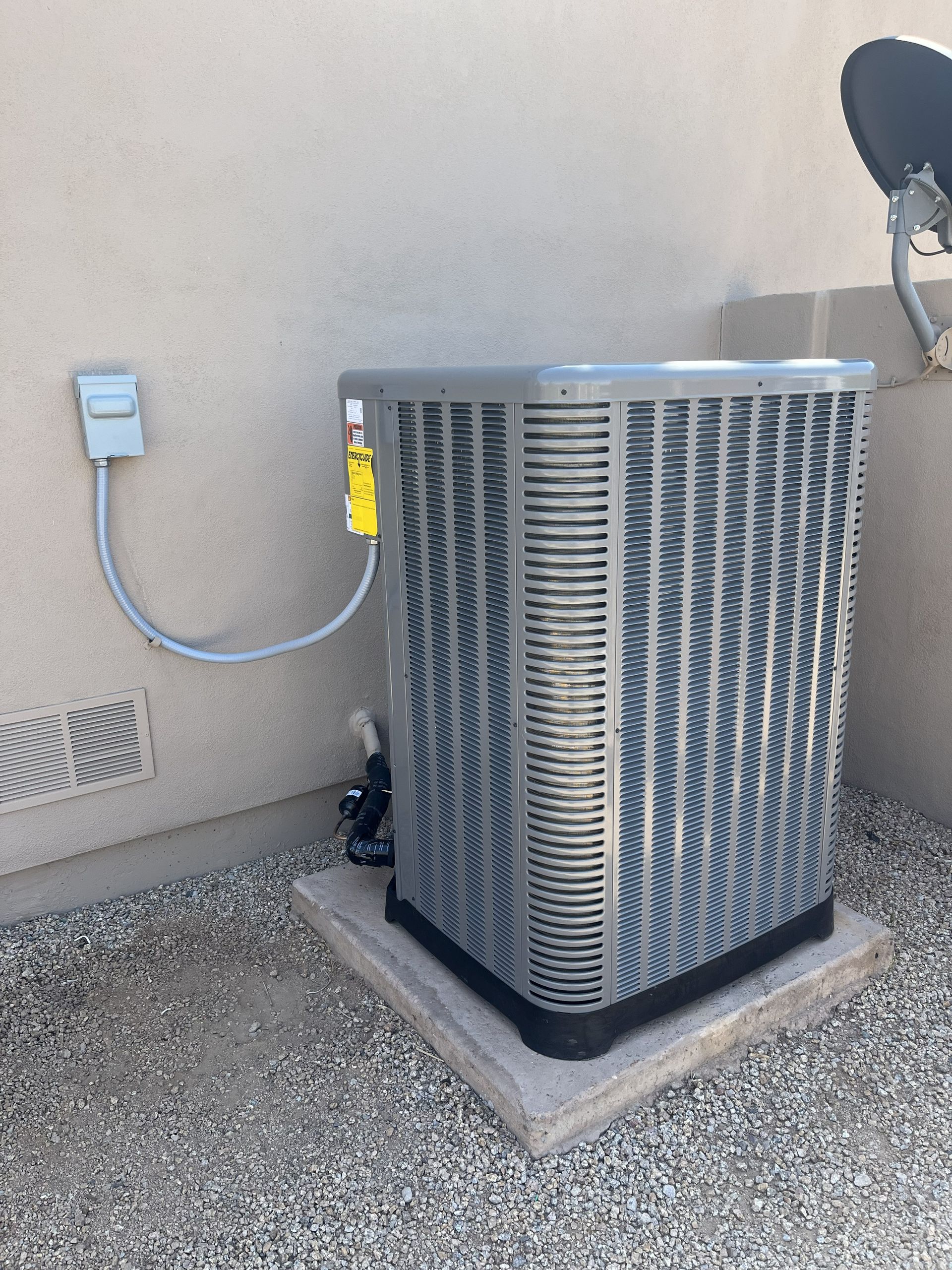 Air conditioning unit on concrete pad next to a beige wall with gray electrical conduit and pebble ground.