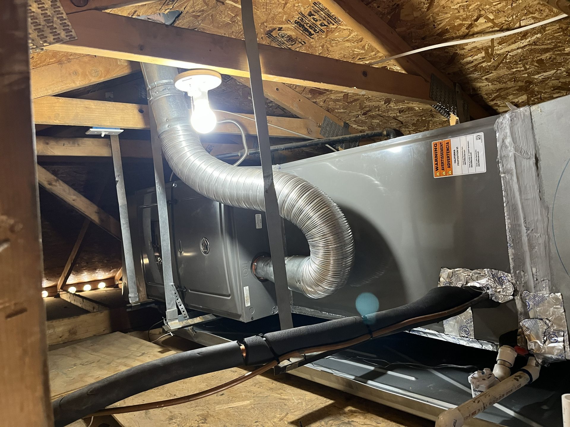 HVAC unit in an attic, with flexible ductwork and light fixture. Wooden beams and insulation visible.