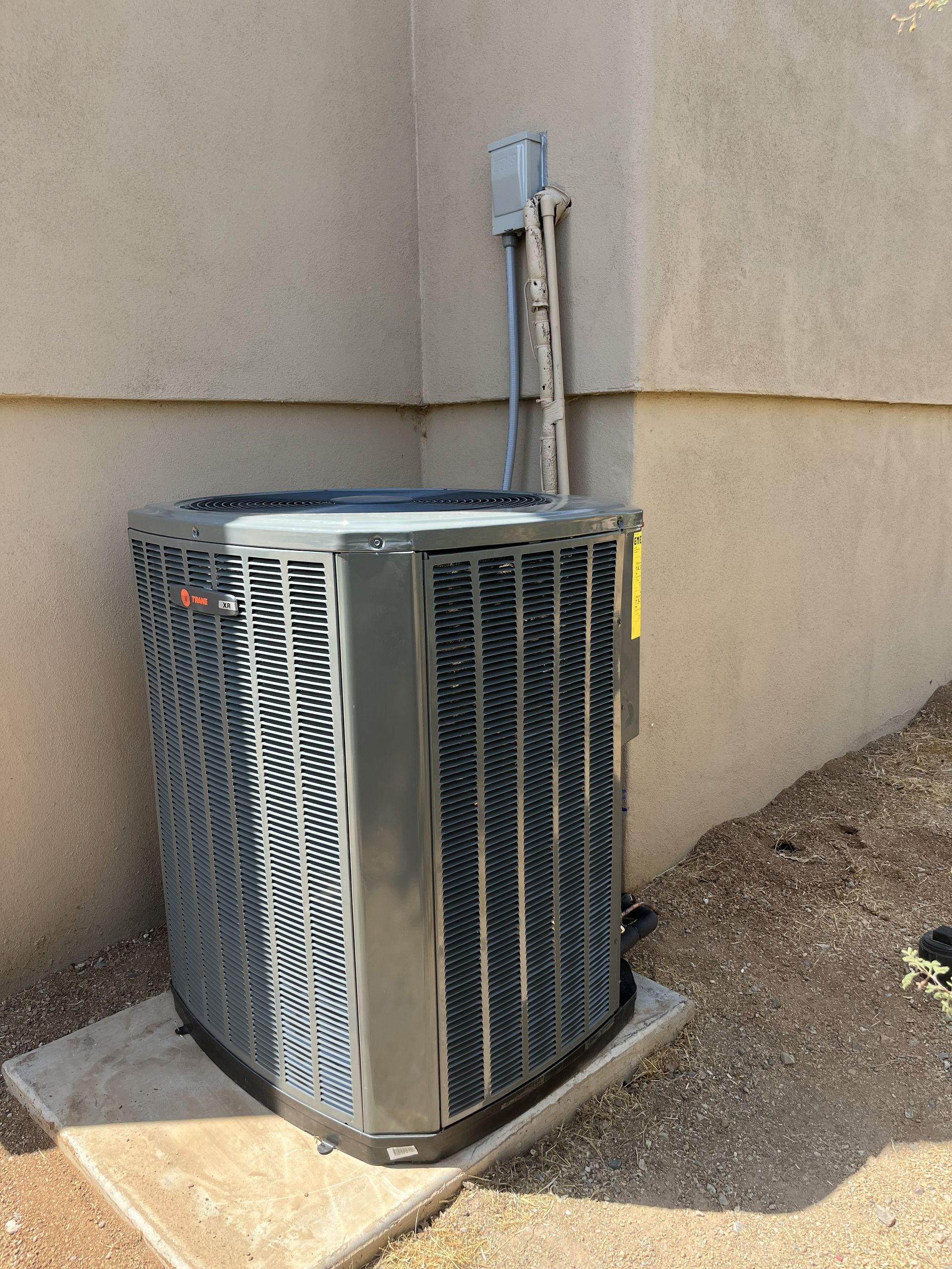 Exterior air conditioning unit against a light brown stucco wall, sitting on a concrete pad.