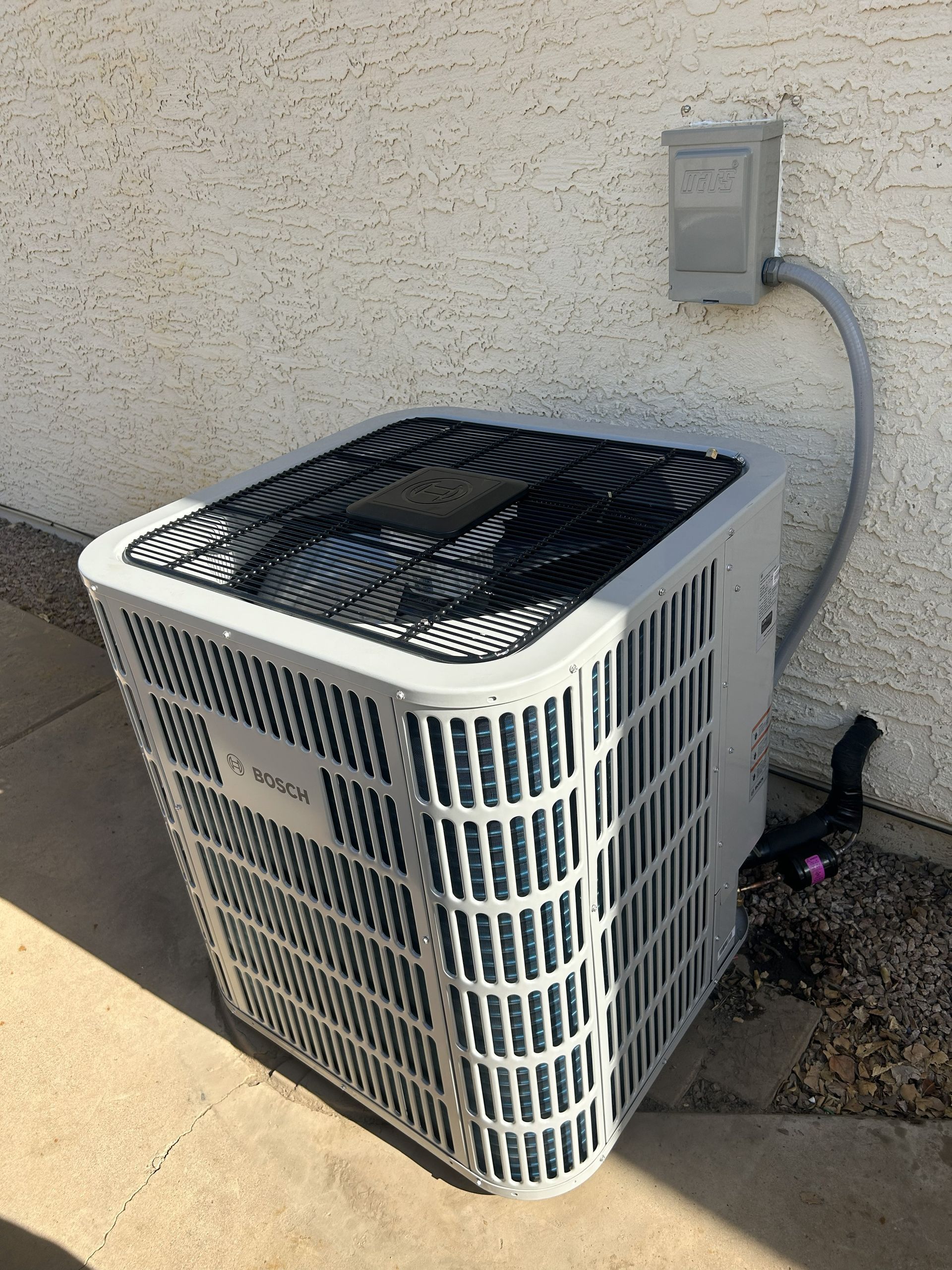 Air conditioner unit outside, mounted against a stucco wall. Gray and white colors. Electrical box above.